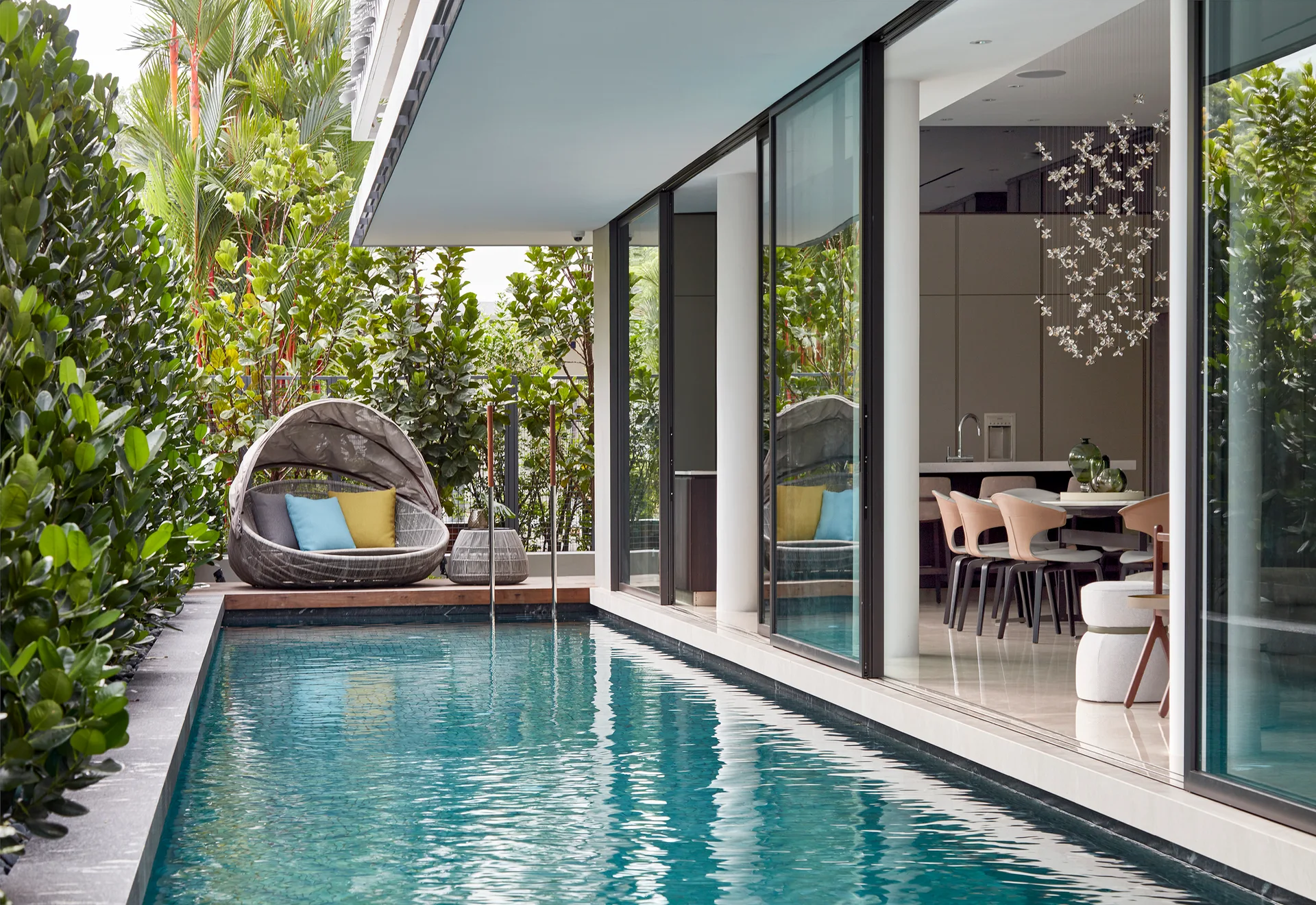 A long swimming pool runs alongside a modern house with open sliding glass doors revealing a dining room, featuring a hooded woven daybed on the deck.