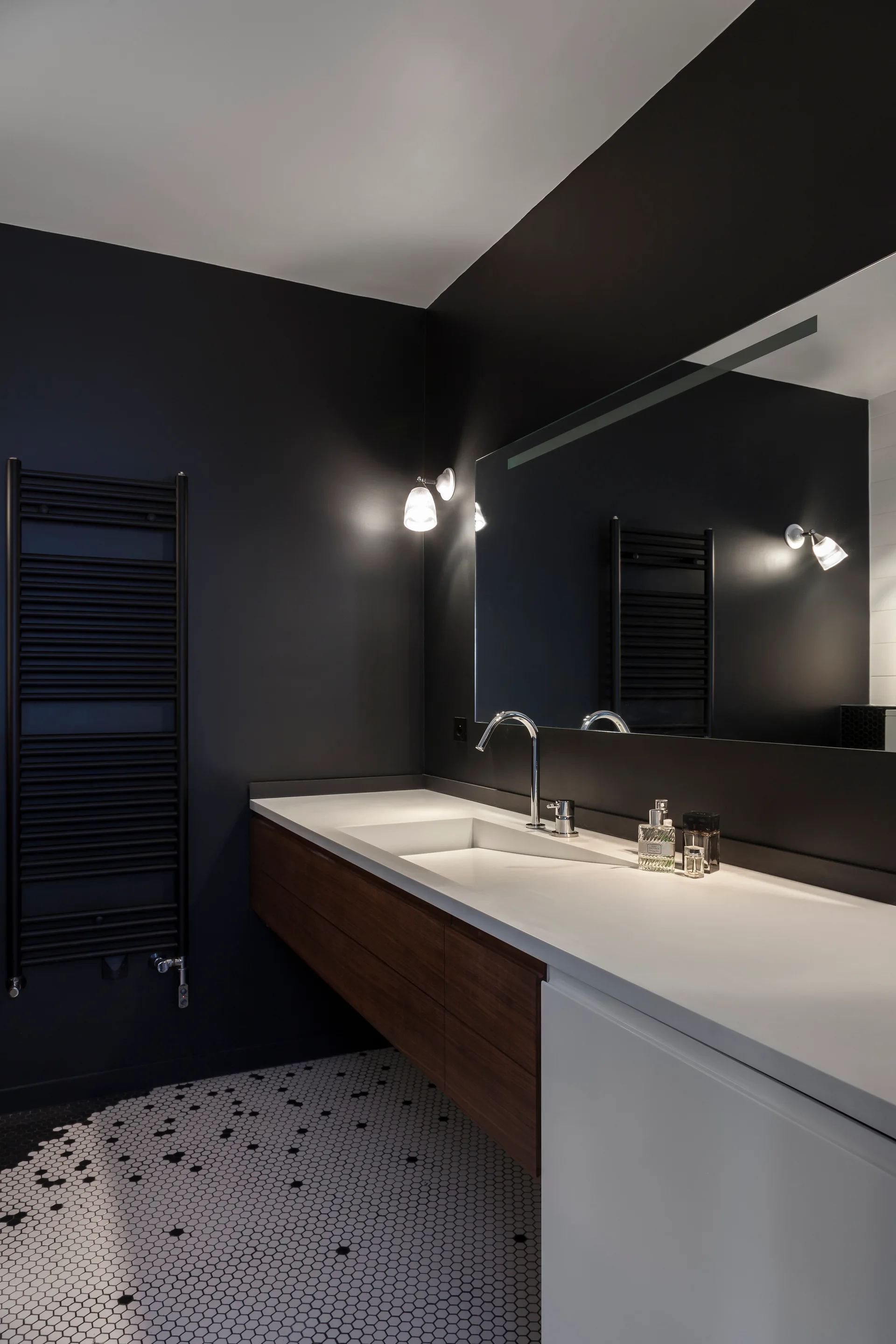 A bathroom with matte charcoal walls features a wooden floating vanity, a large frameless mirror, and geometric black and white floor tiles.