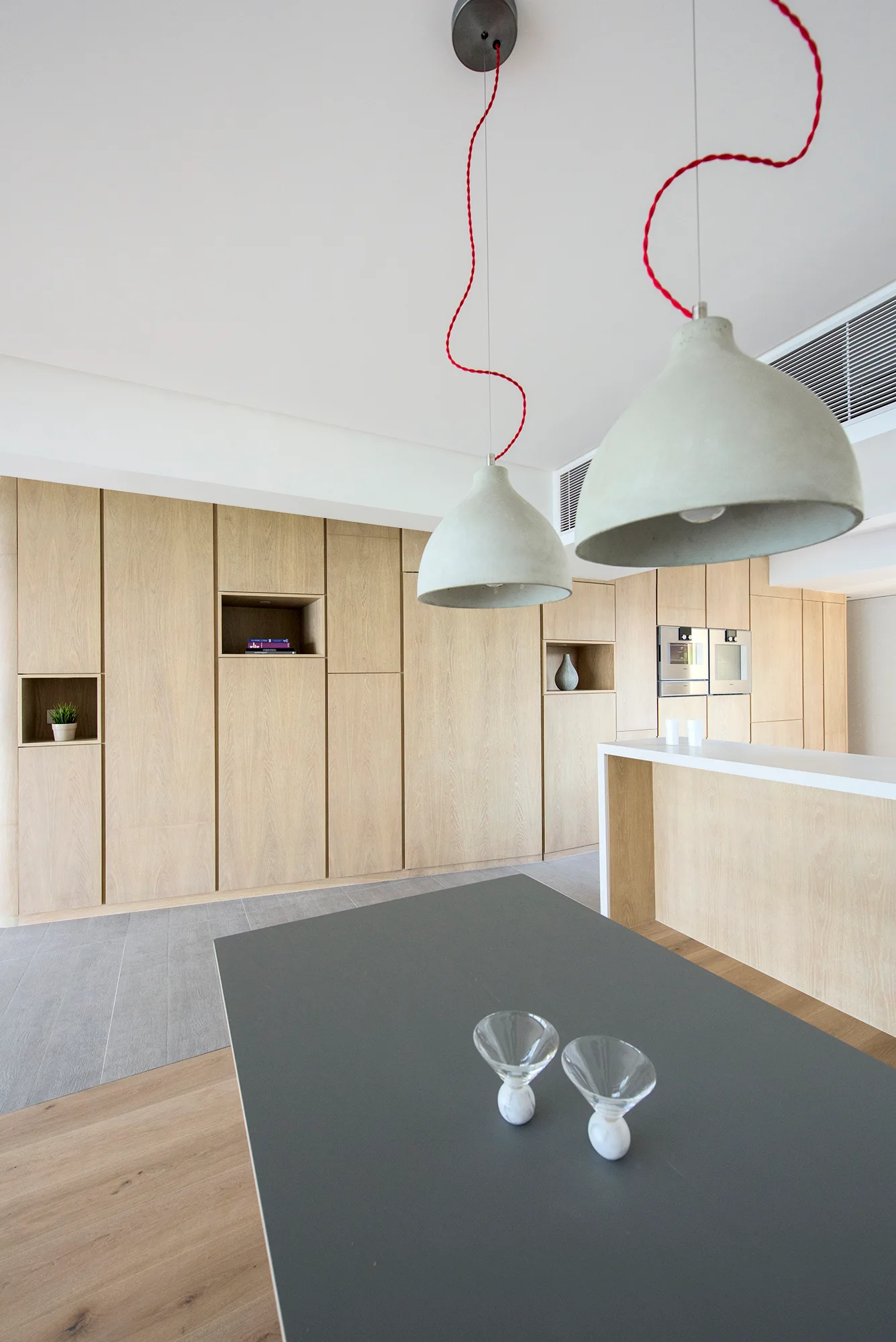A modern kitchen featuring full-height light wood cabinetry with recessed niches, concrete pendant lights with wavy red cords, and a grey island.