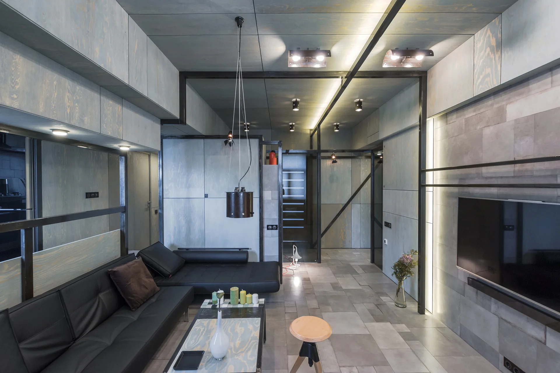A modern living room with grey plywood paneling on the walls and ceiling, a black leather sectional sofa, and black metal structural framing.