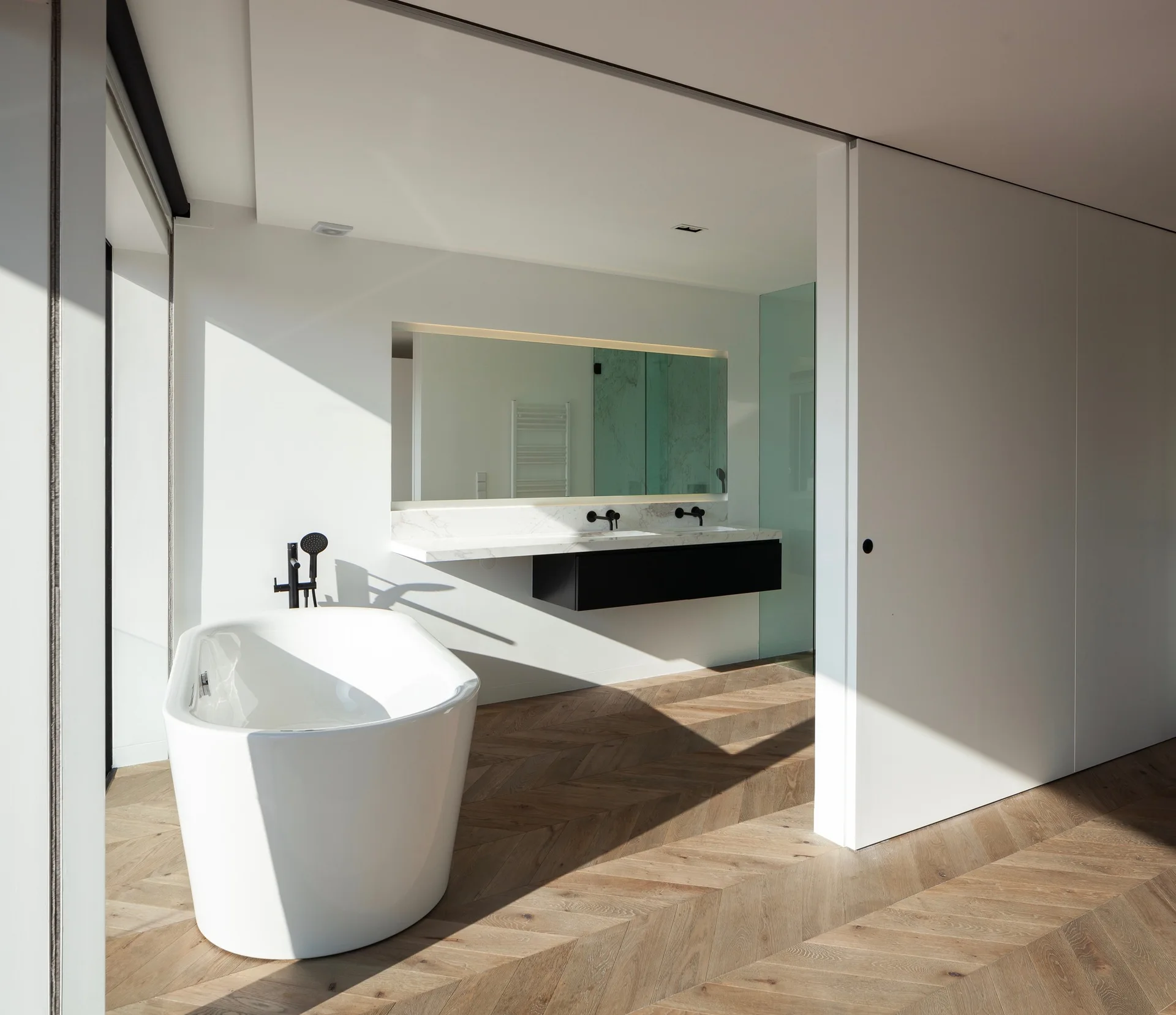 A minimalist white bathroom features a freestanding bathtub, a floating black and marble vanity, and warm wood herringbone flooring.