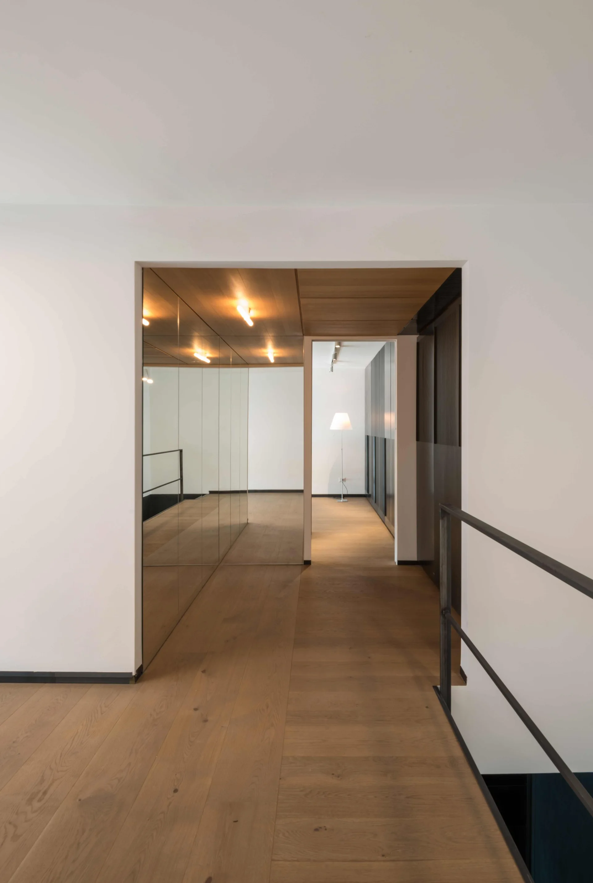 A minimalist hallway featuring light wood flooring, a wood-paneled ceiling insert, a large mirrored wall, and a black metal railing.