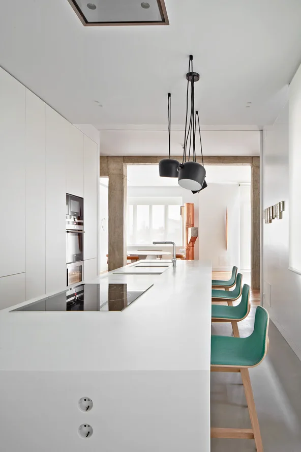 A modern white kitchen features a long minimalist island with green bar stools, flat-panel cabinets, and black pendant lights.