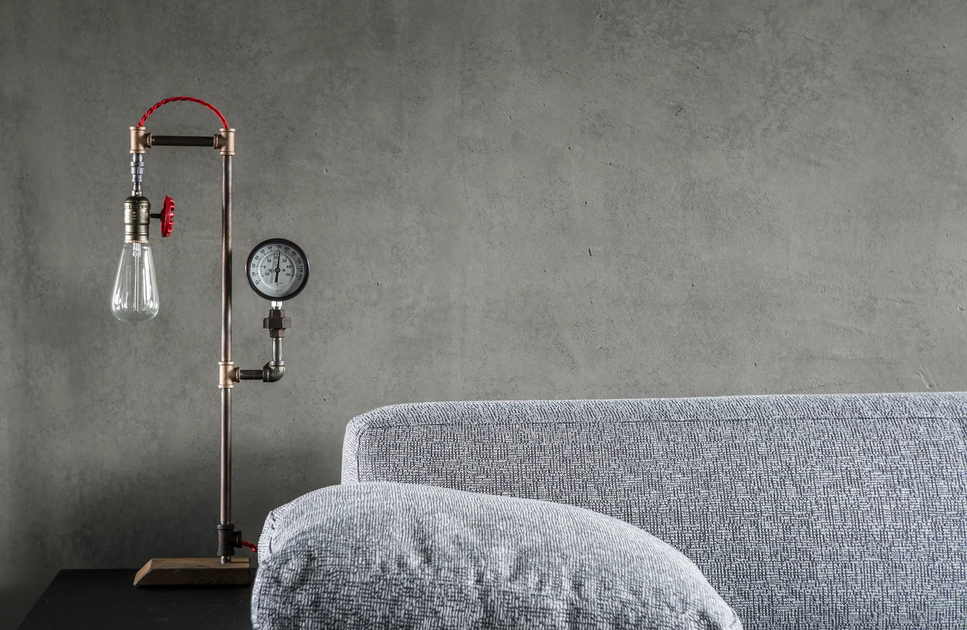 An industrial table lamp made of metal pipes sits next to a grey fabric sofa in front of a raw concrete wall.
