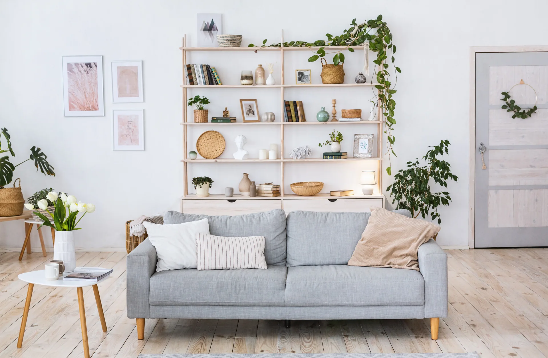A light grey sofa sits in front of a tall wooden open shelving unit filled with books, plants, and decorative objects on a light wood floor.