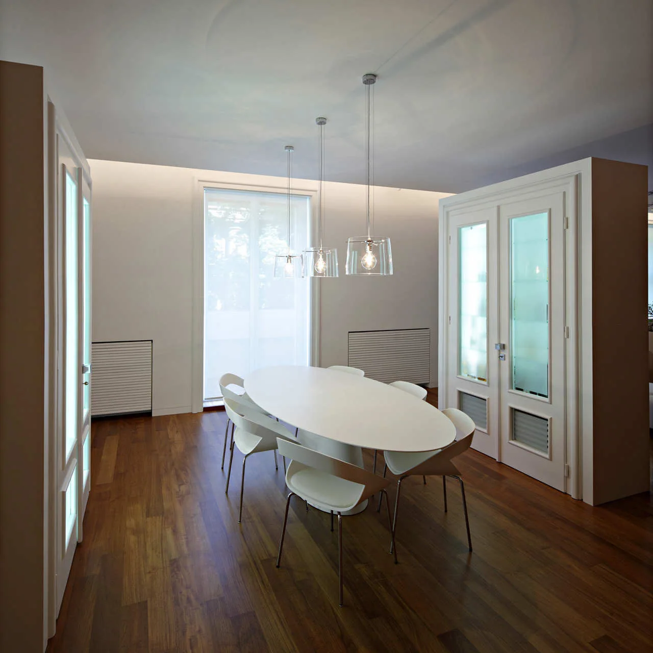 A minimalist dining room featuring a central white oval dining table, matching chairs, dark wood floors, and three glass pendant lights.