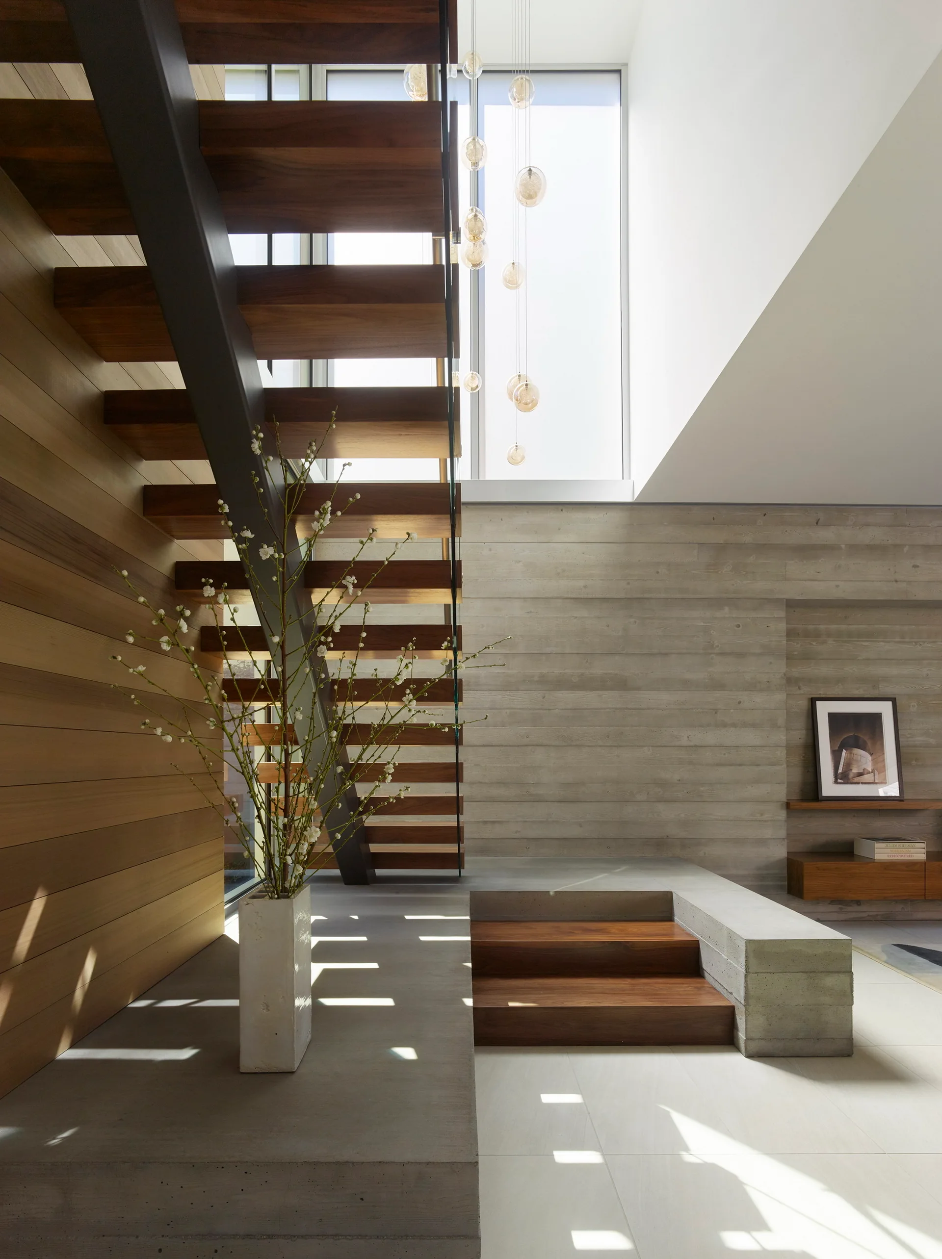 A modern open-riser wooden staircase with a metal support sits beside a board-formed concrete wall and a tall vase with white blossoms.