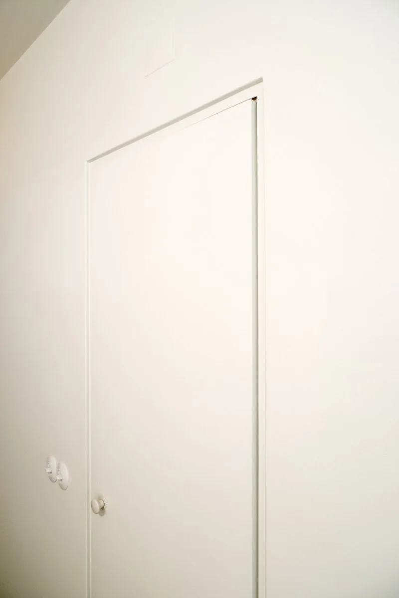A smooth white wall features a minimalist flush-mount door with a matching white knob and round ceramic light switches.