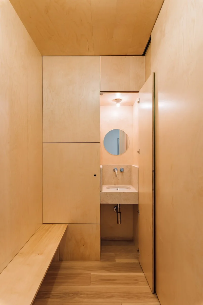 A minimalist powder room featuring walls clad in light plywood panels, a built-in wooden bench, a circular mirror, and a floating stone sink basin.