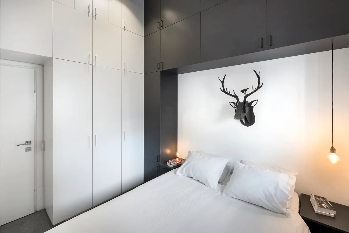 A modern bedroom features a white bed framed by asymmetrical black and white built-in cabinets and a black stag head sculpture on the wall.