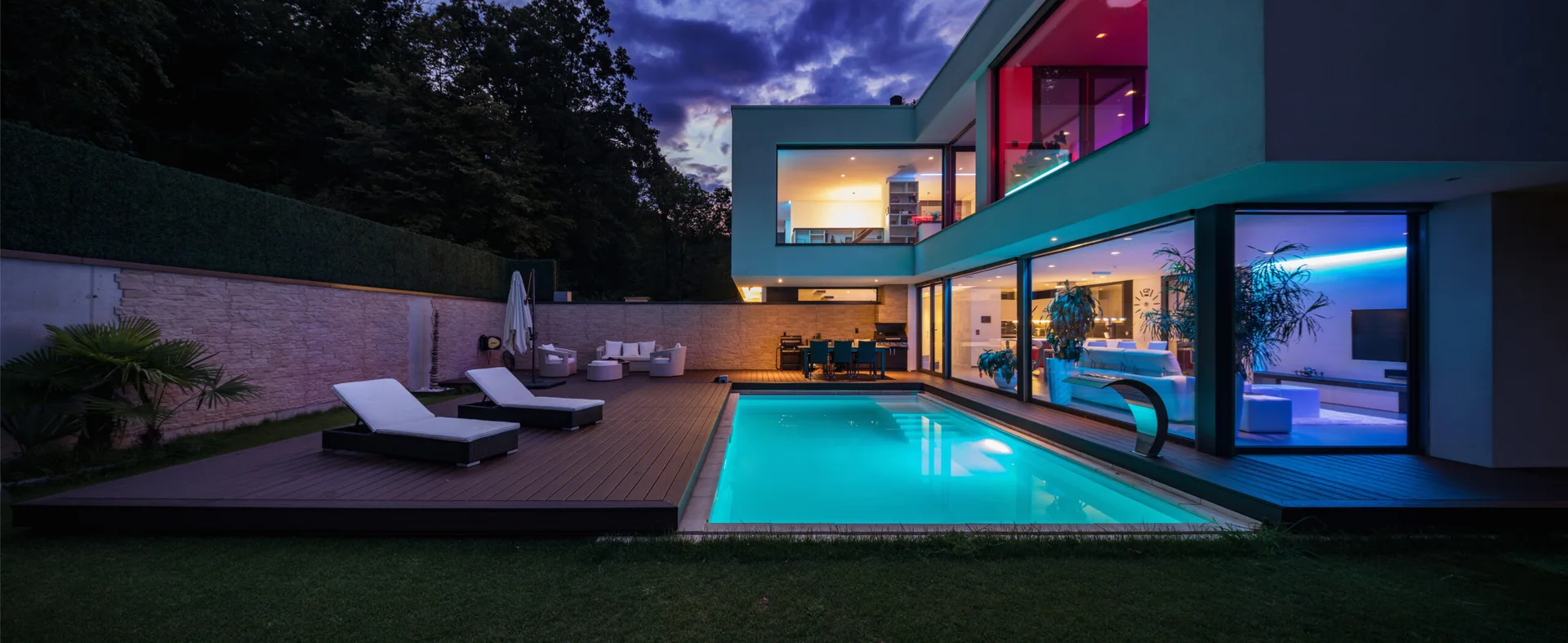 A modern two-story house at twilight features a bright swimming pool, dark wood deck, and colorful interior lights visible through large windows.