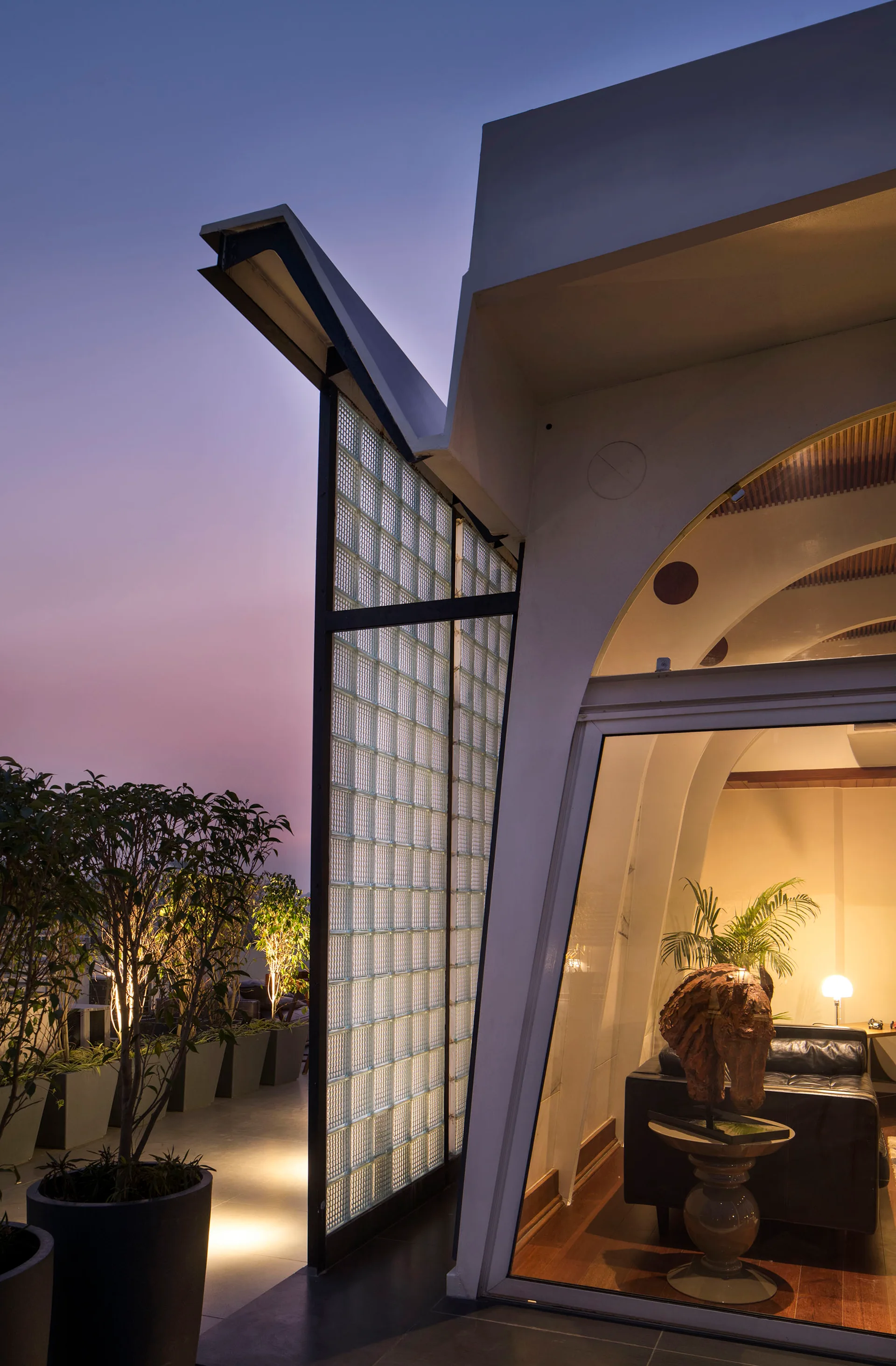 A modern terrace at dusk featuring a translucent glass block partition, potted plants with up-lighting, and a view into a warm interior.