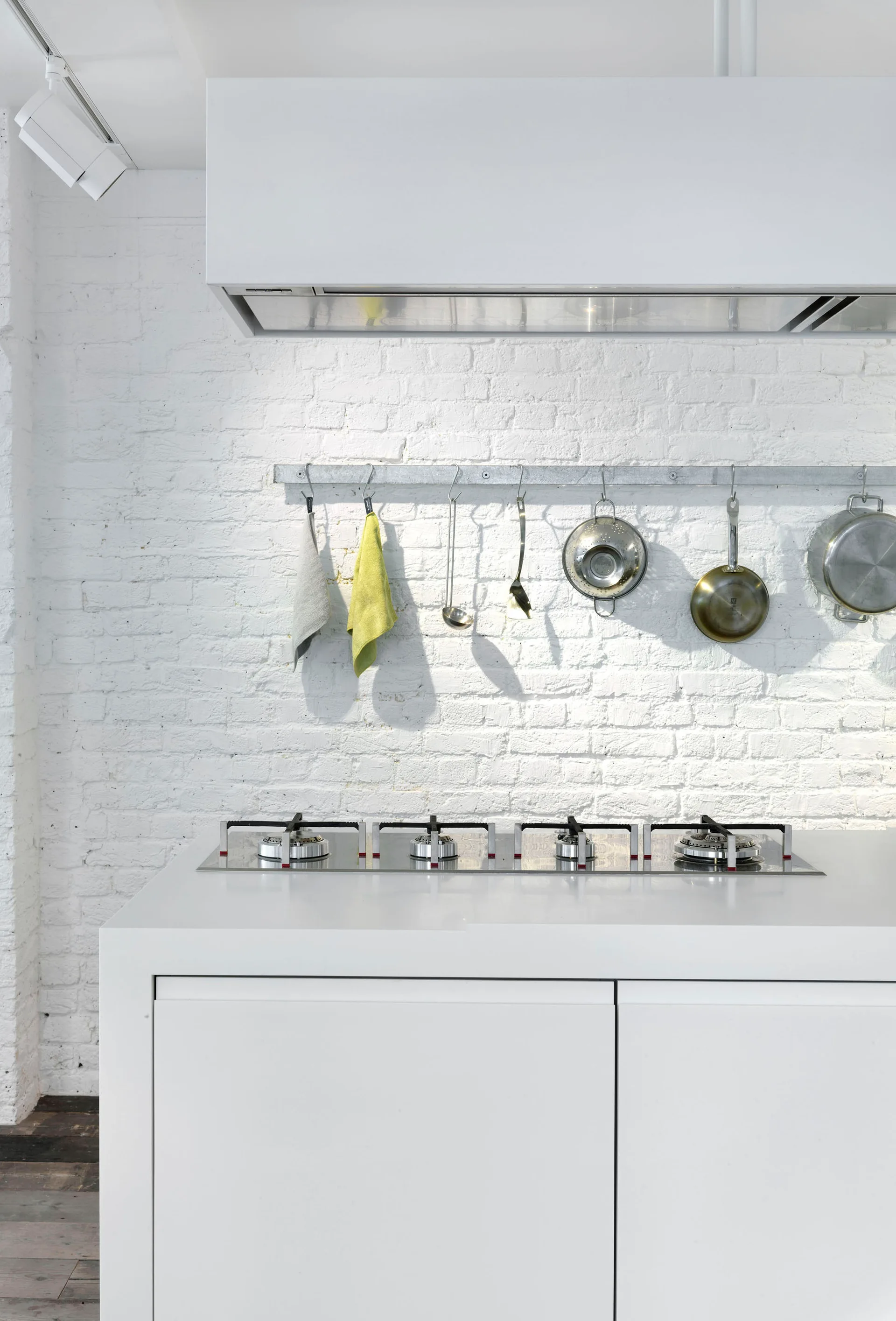 A minimalist white kitchen island with a gas cooktop is set against a white painted brick wall featuring a hanging metal utensil rail.