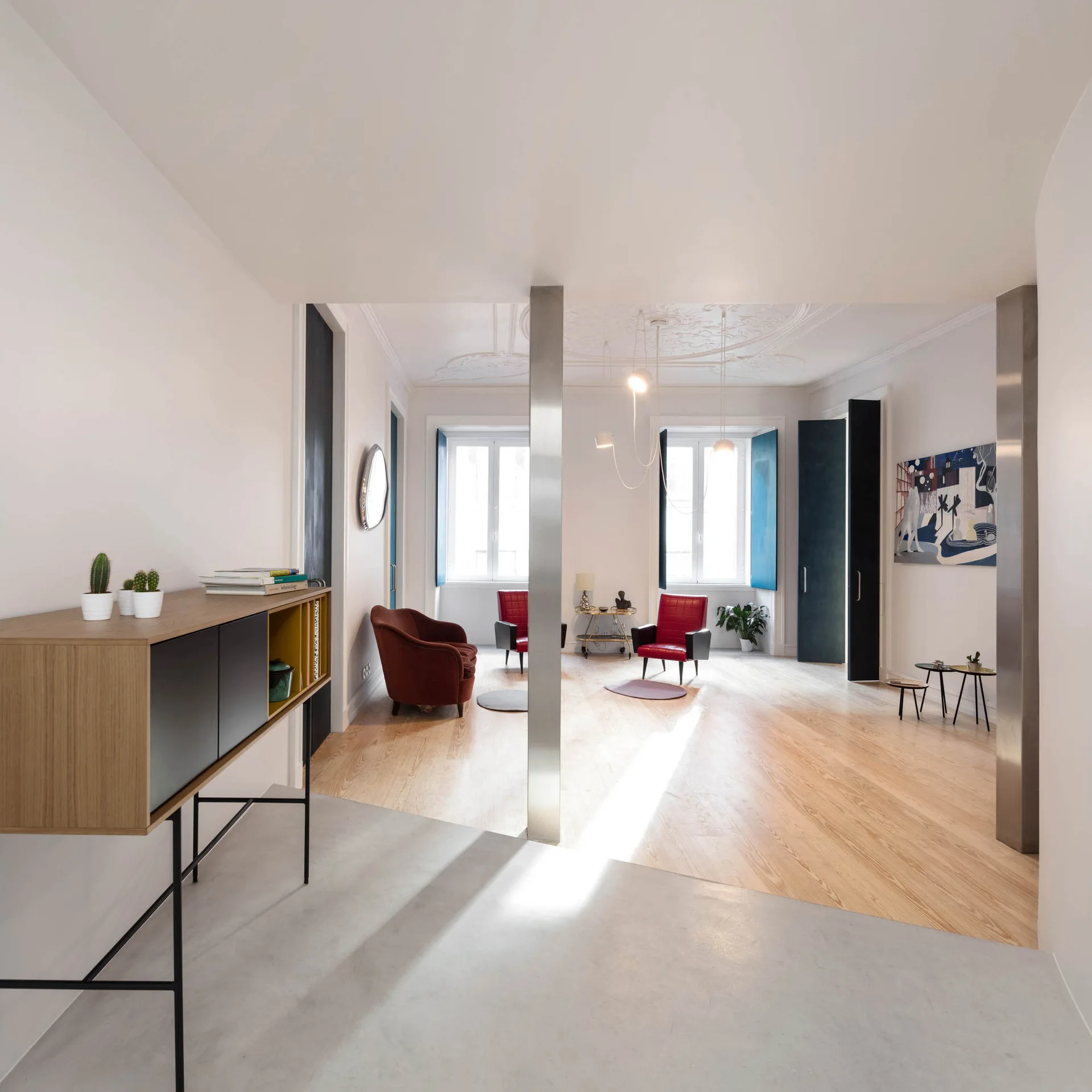 A modern white living room with polished steel columns, light wood flooring, red armchairs, and an ornate plaster ceiling.