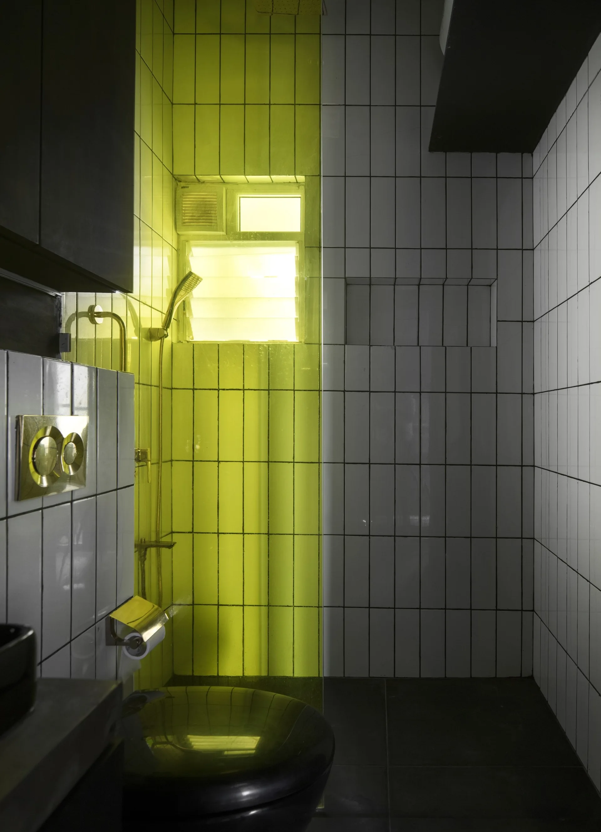 A modern bathroom featuring white vertical tiles, a translucent yellow glass shower partition, a black toilet, and gold metallic fixtures.