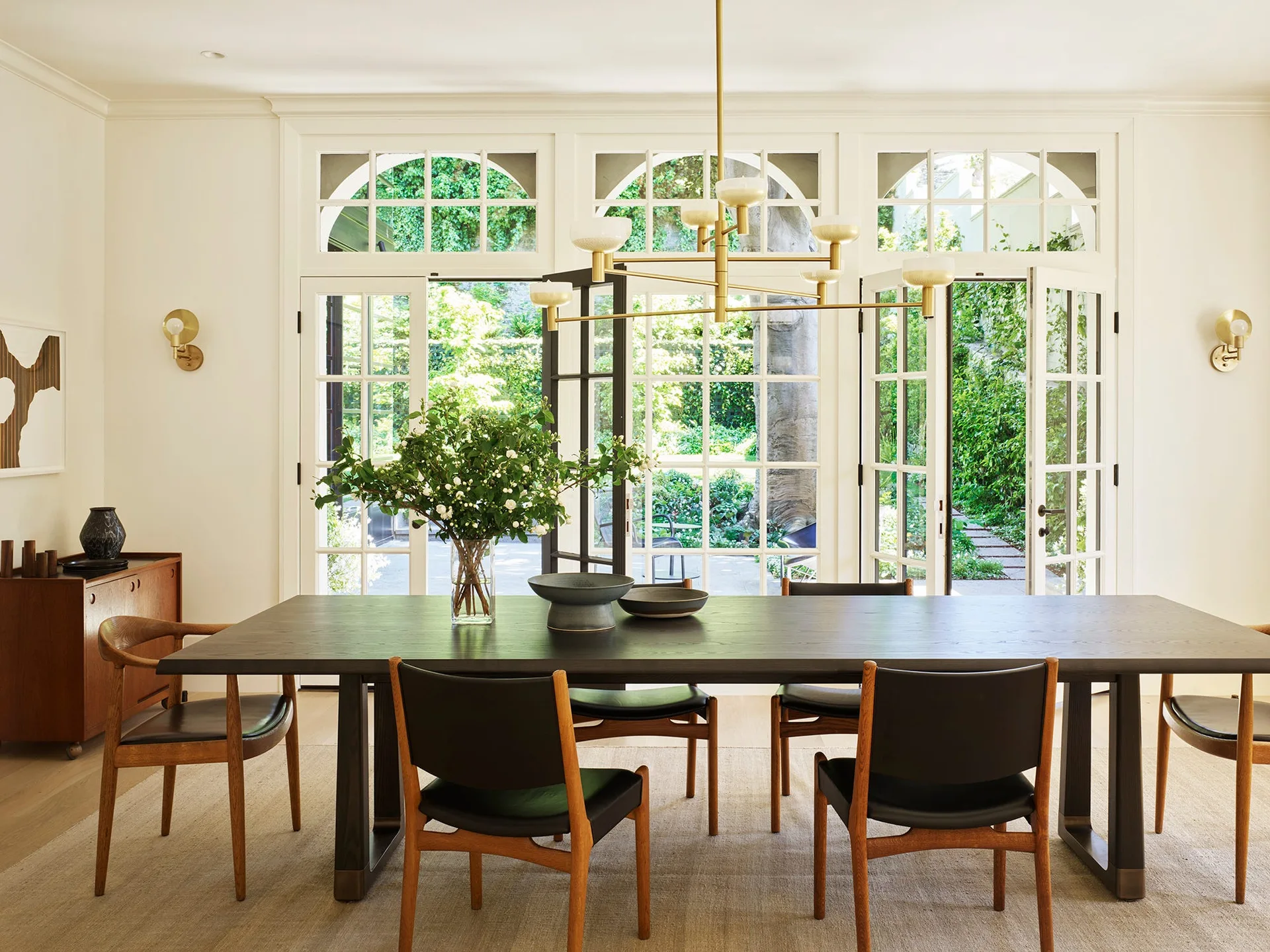 A dark wood dining table and leather chairs sit on a woven rug before white-framed French doors opening onto a lush garden.