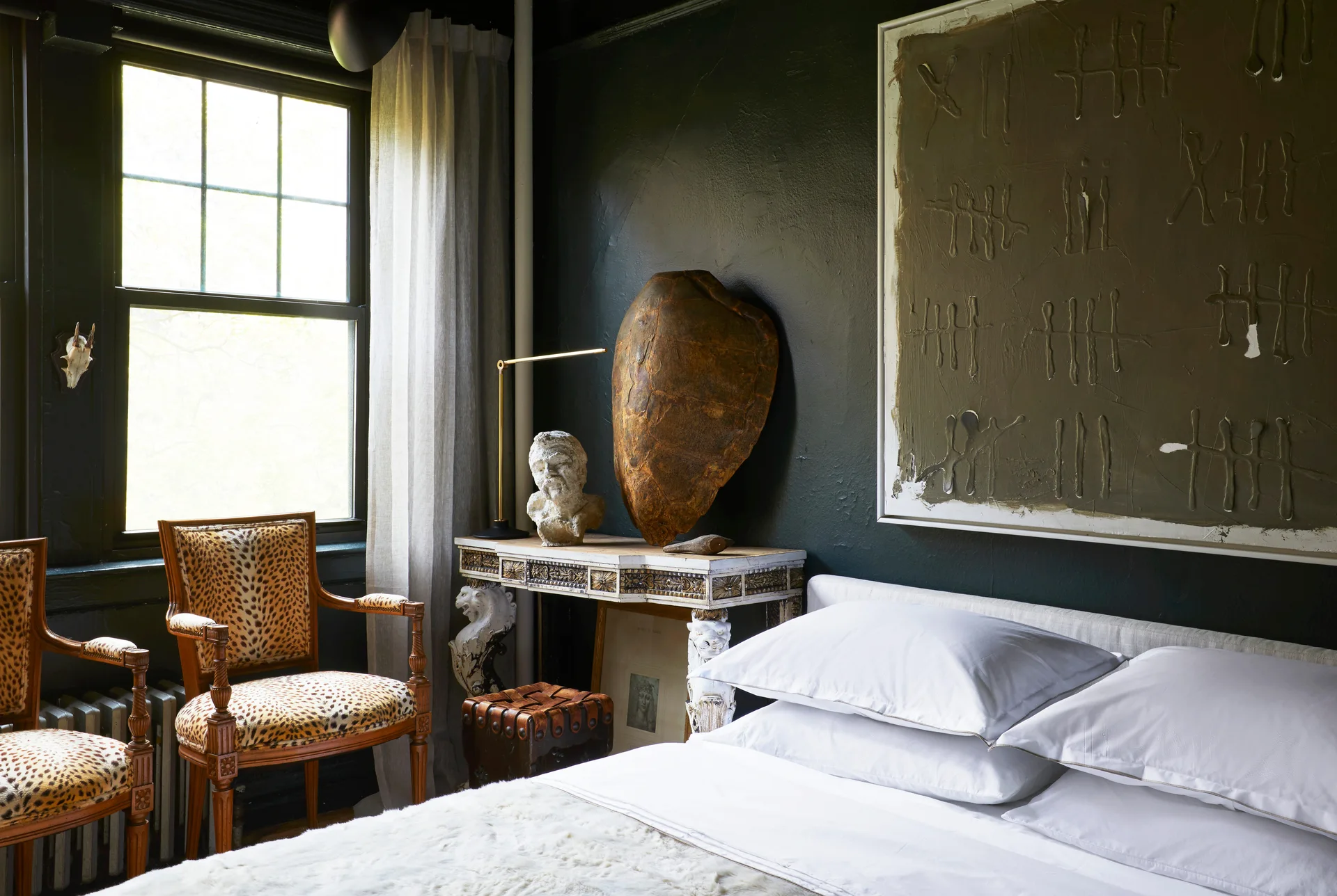 A moody bedroom features dark charcoal walls, a white bed, leopard-print armchairs near a window, and an ornate console table with sculptures.