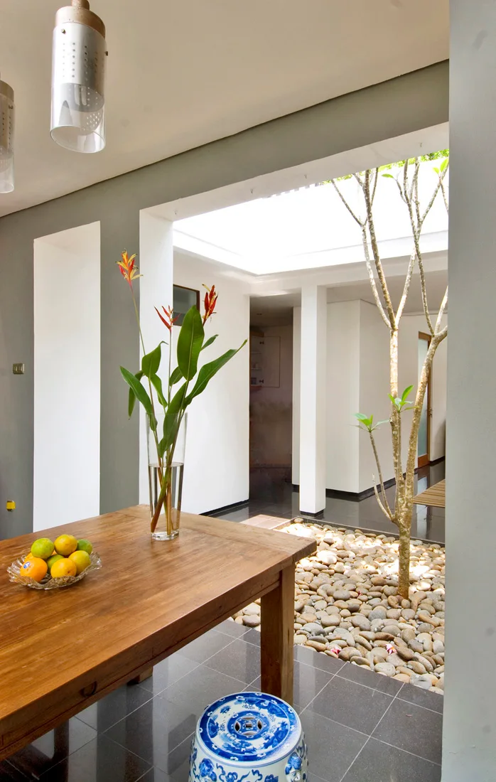 A wooden dining table sits next to a large opening looking into an indoor courtyard filled with light river stones and a small tree.