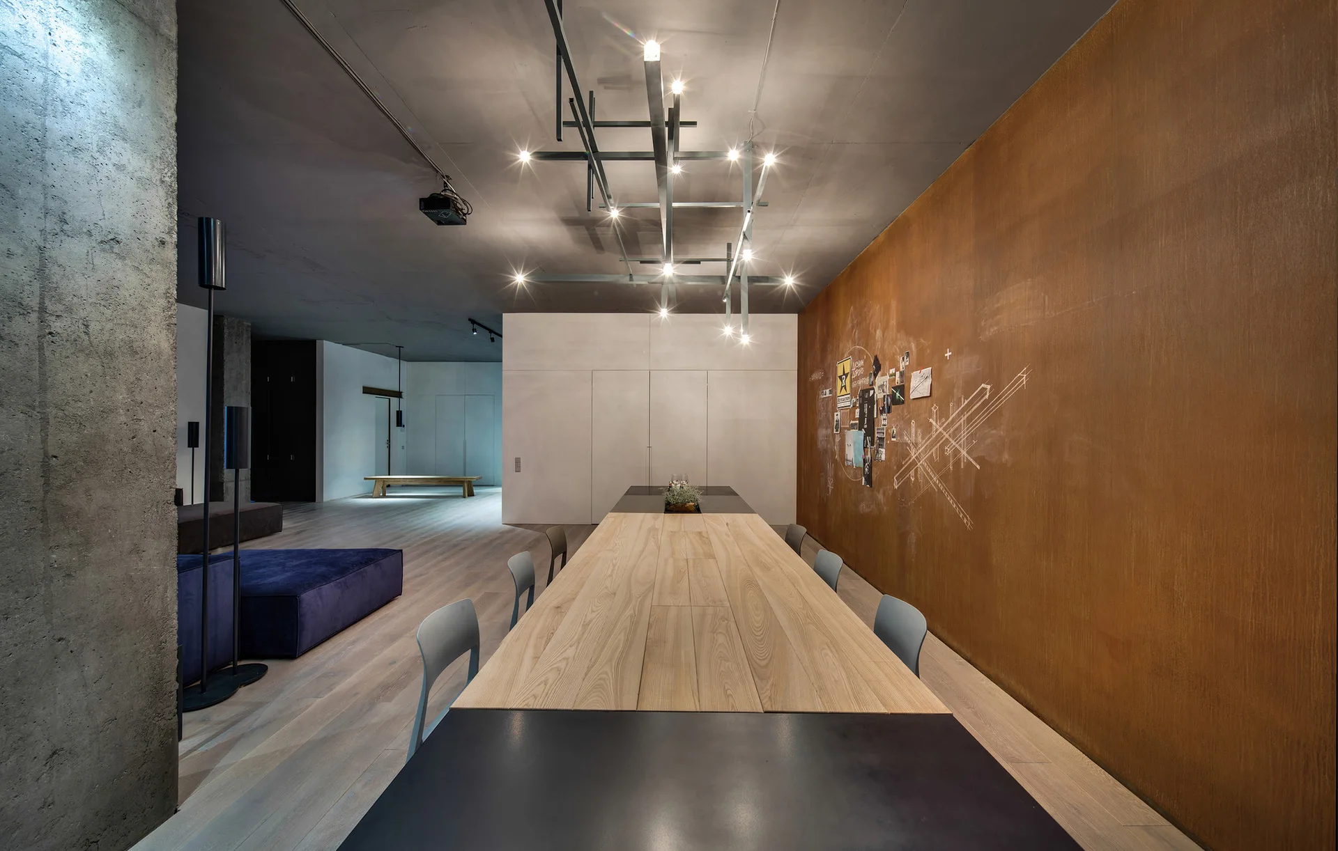 A long dining table with a wood and metal top stands under a geometric grid light fixture next to a large rust-colored accent wall.