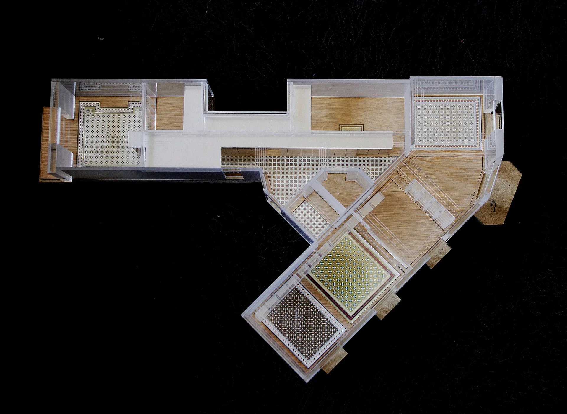 An overhead view of a physical architectural scale model featuring transparent acrylic walls, warm wood flooring, and varied geometric tile patterns.