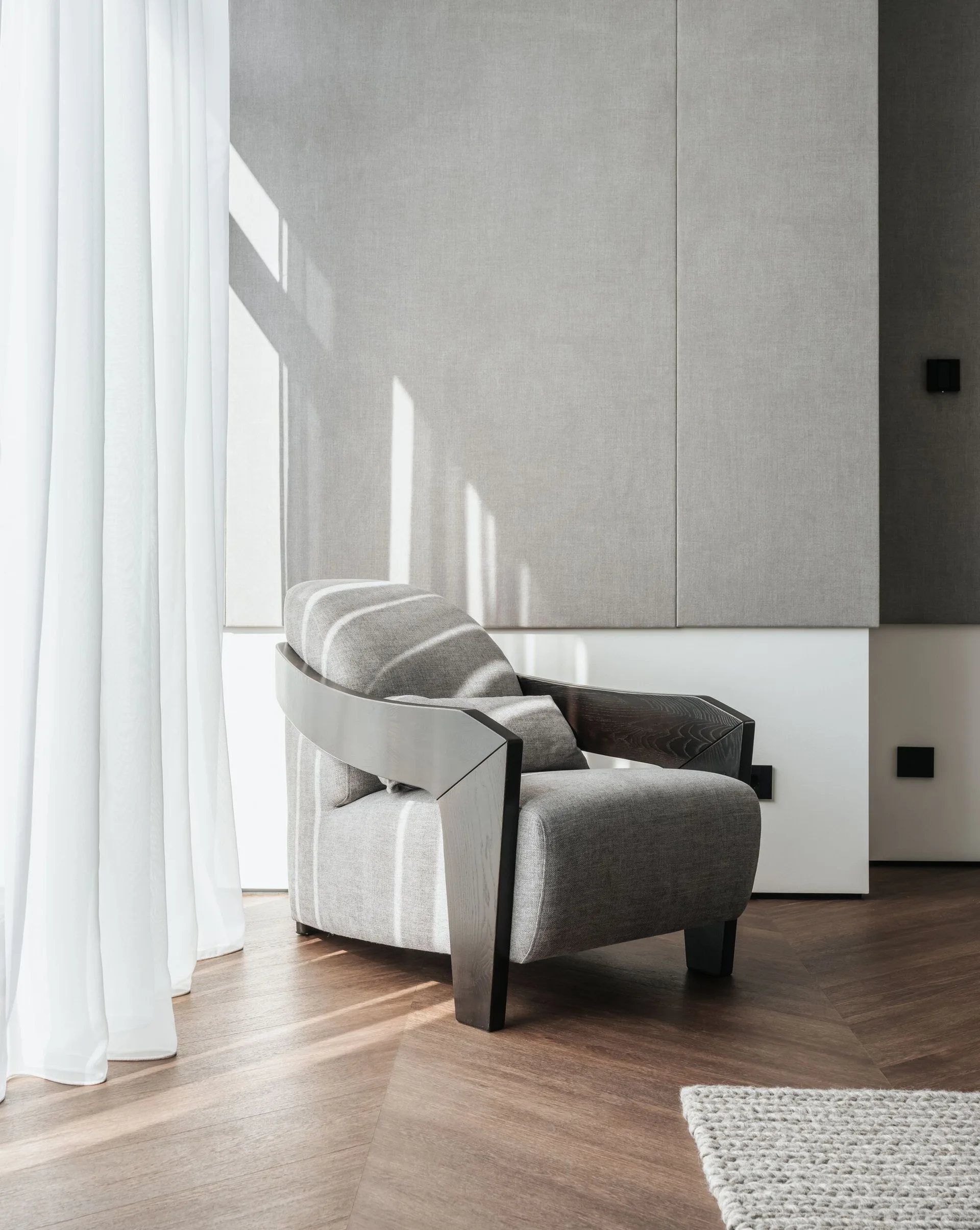 A grey upholstered armchair with a dark wood frame stands on chevron wood flooring next to a sheer white curtain.