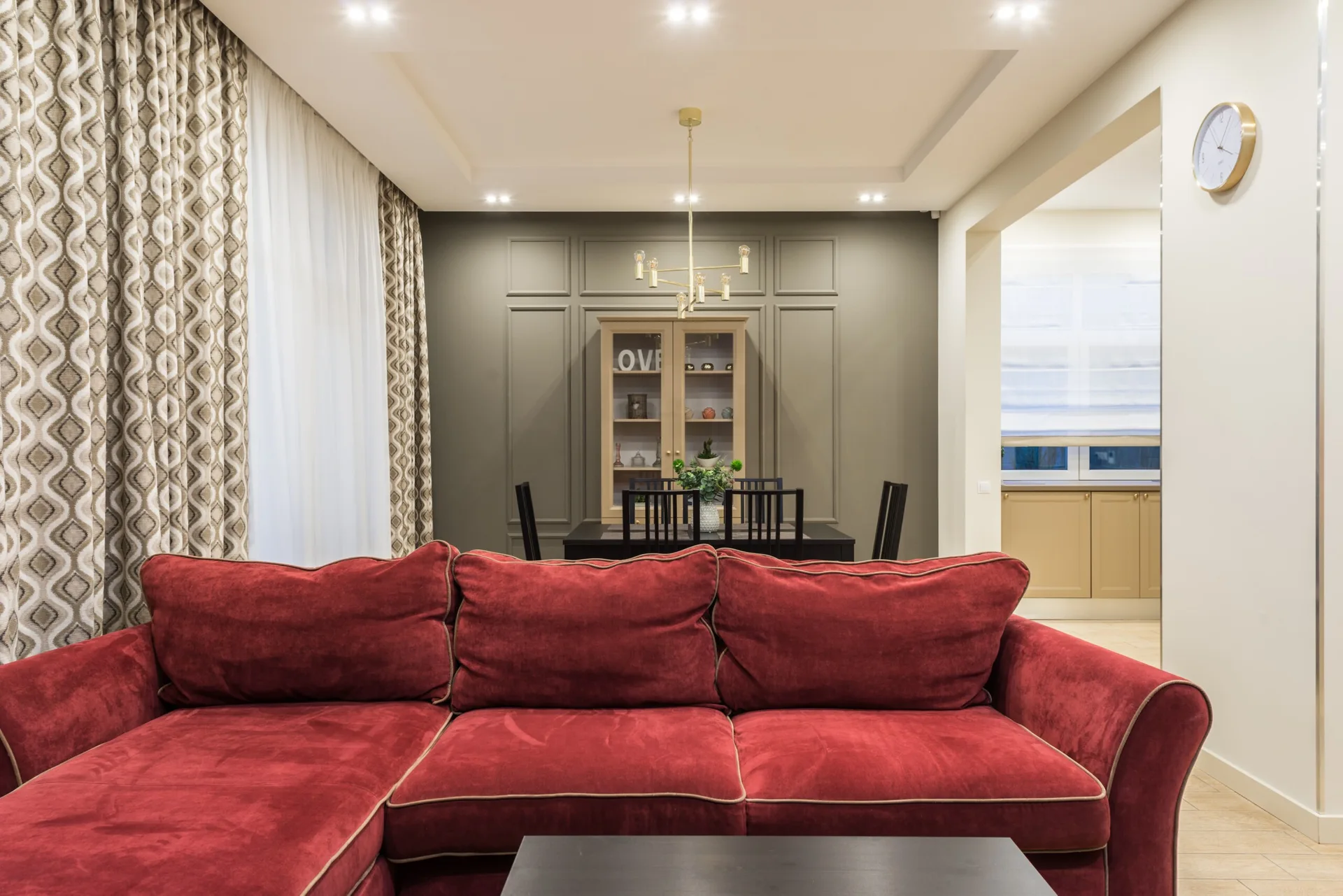 A living room featuring a plush red velvet sectional in the foreground, looking toward a dining table, gold chandelier, and a grey paneled accent wall.