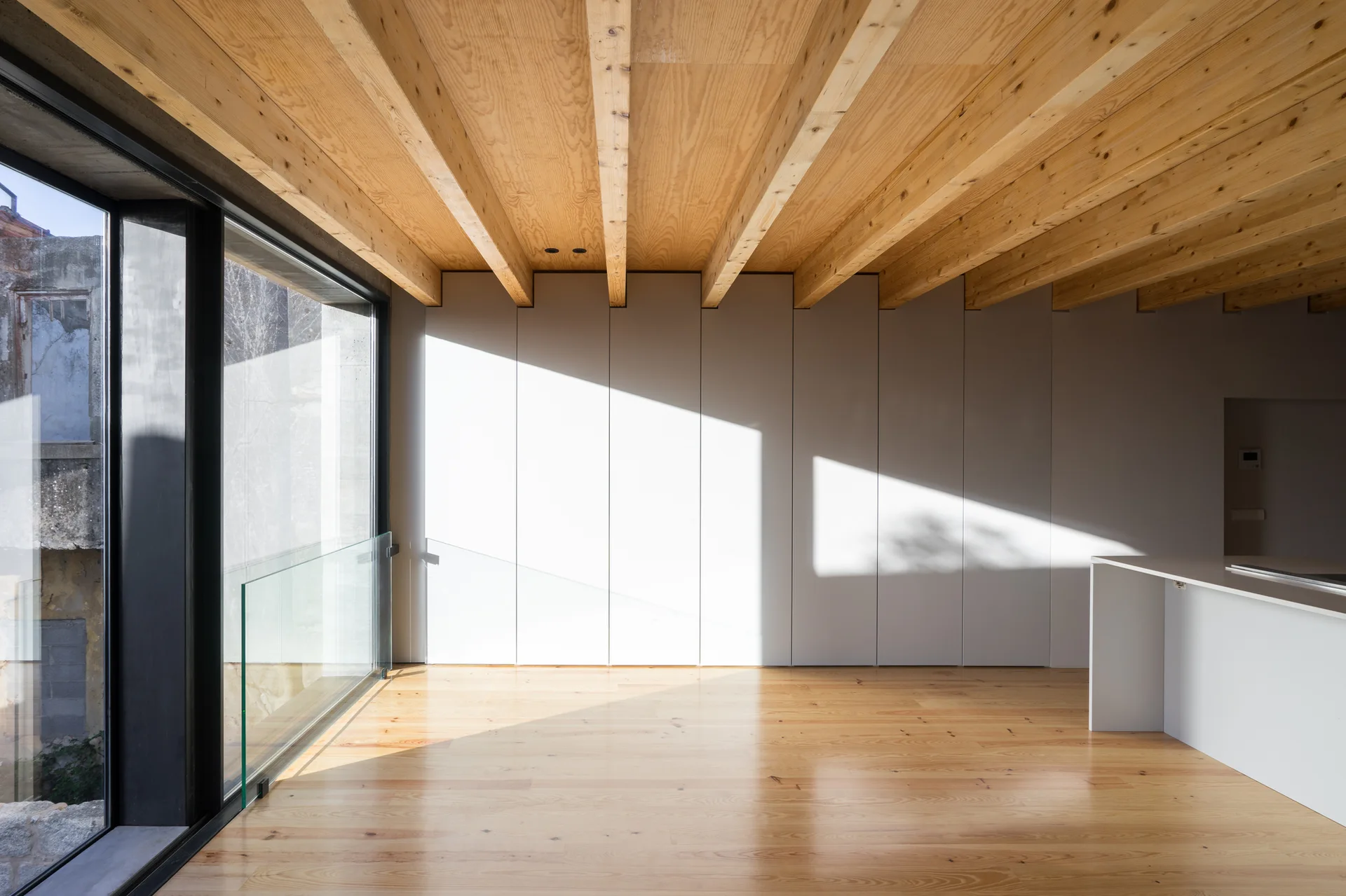 A minimalist interior room featuring exposed light wood ceiling beams, matching hardwood floors, a seamless white cabinetry wall, and a large window.