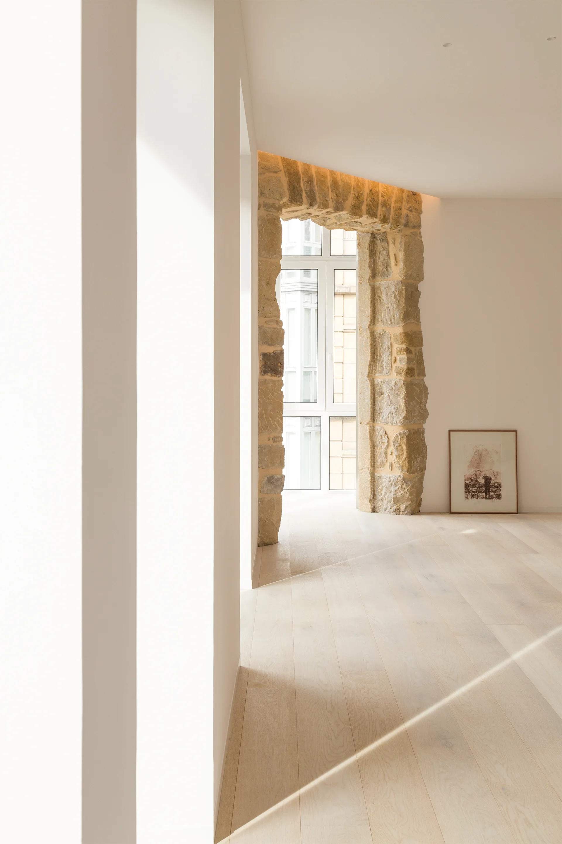 A minimalist room featuring a rough stone archway around a tall window, smooth off-white walls, blonde wood floors, and a framed picture on the floor.