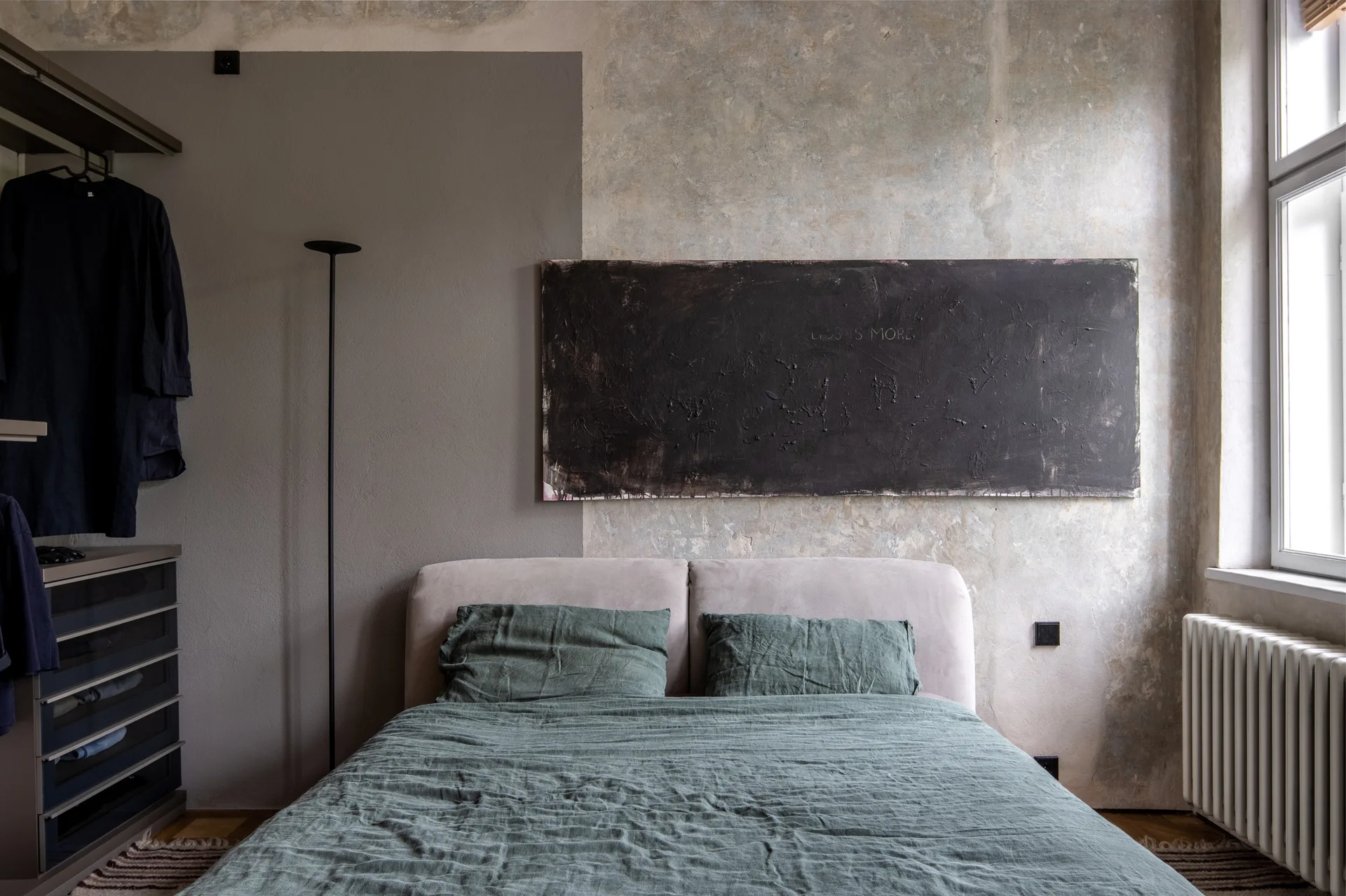 A bed with green linen bedding sits against a two-tone gray and concrete wall with a large black textured canvas hanging above the headboard.