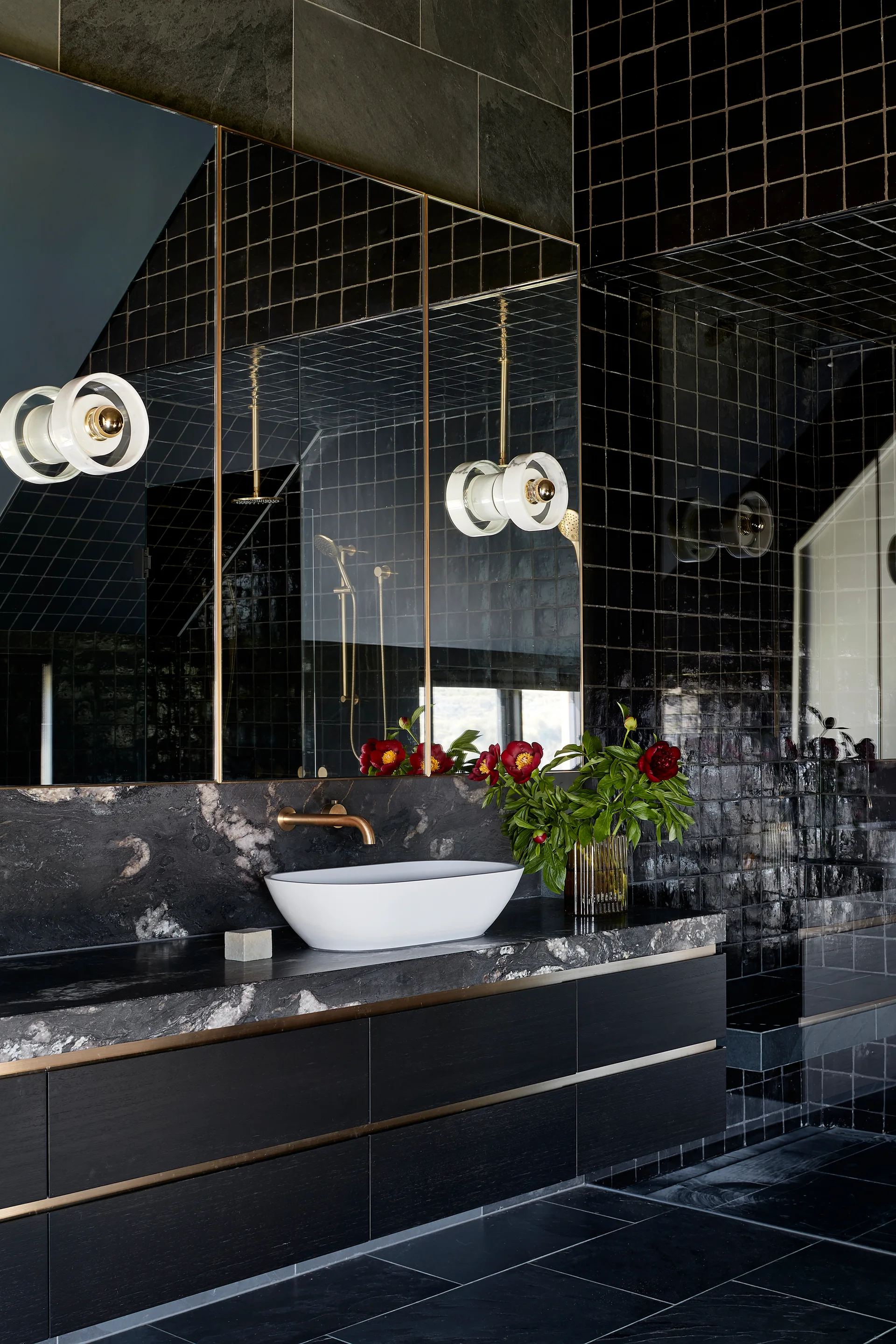 A modern bathroom features a dark wood floating vanity with a white vessel sink, black marble countertop, and glossy black square tiled walls.