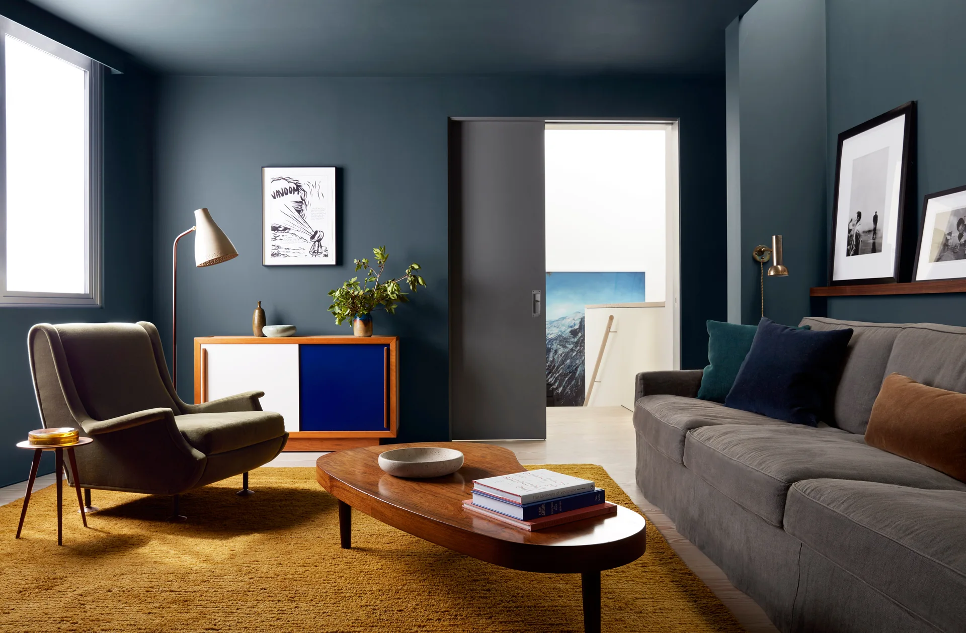 A living room features deep blue-grey walls and ceiling, a dark grey sofa, a textured mustard yellow rug, and mid-century modern wood furniture.