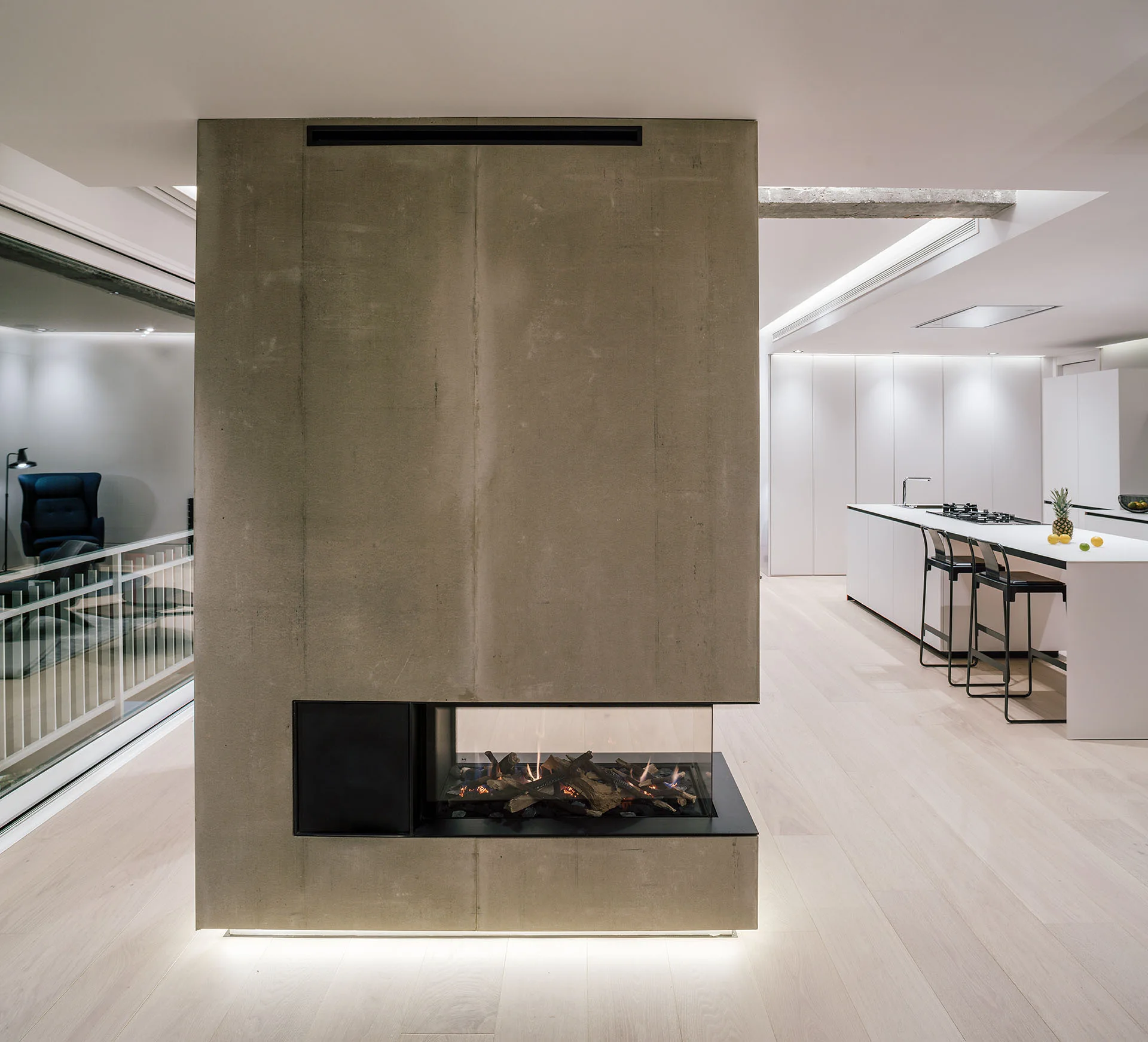 A minimalist open-plan living room with a large central concrete fireplace dividing the space from a white modern kitchen.