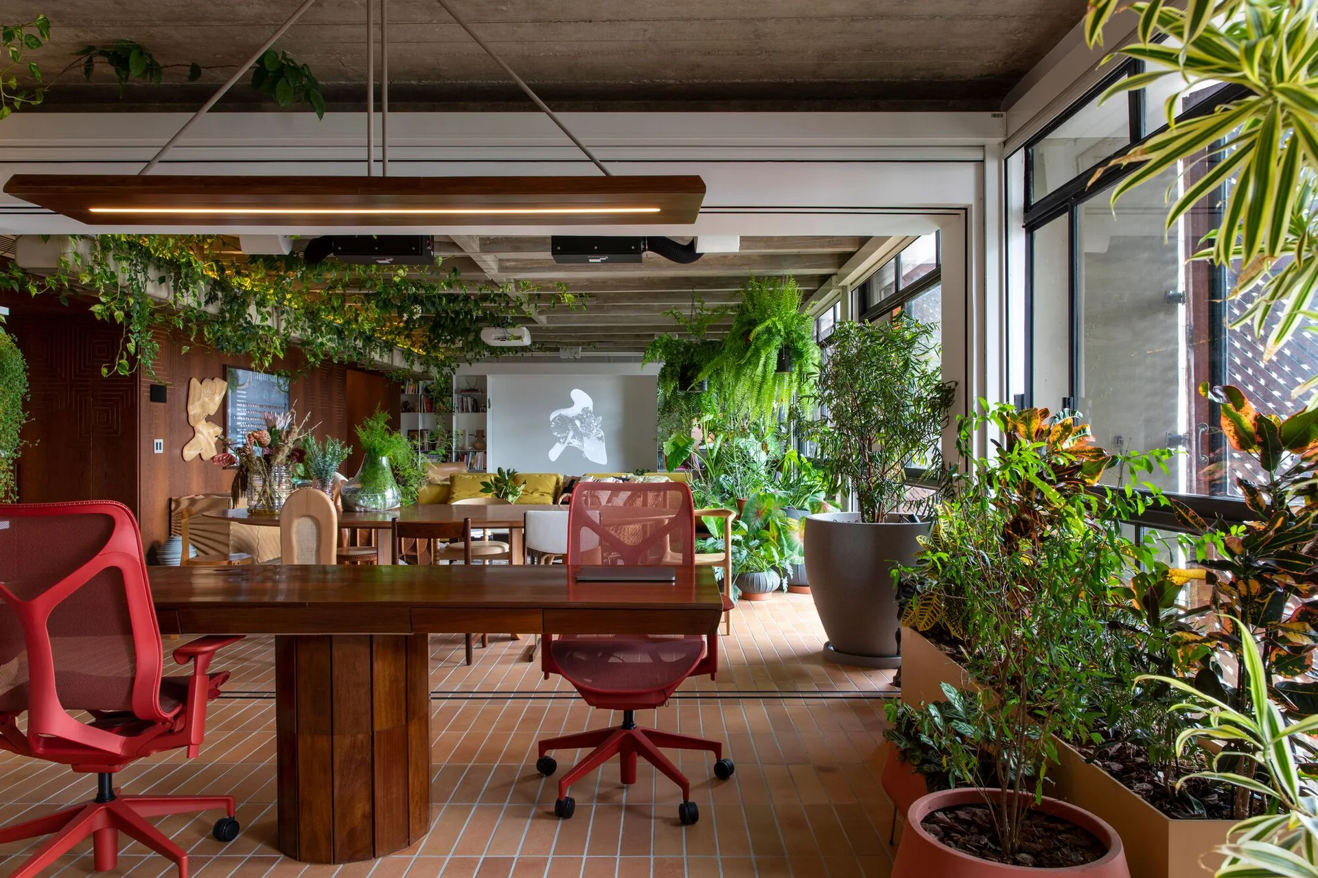 A biophilic office features a wood desk with red mesh chairs, a terracotta tiled floor, and numerous indoor plants under an exposed concrete ceiling.