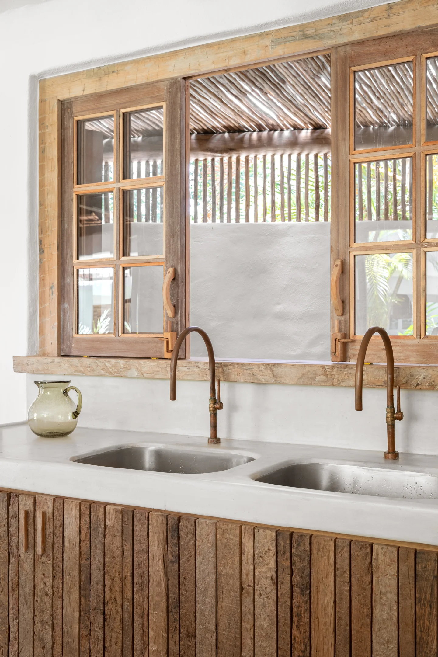A rustic double vanity with stainless steel sinks and bronze faucets set in a plaster countertop below a weathered wooden window.