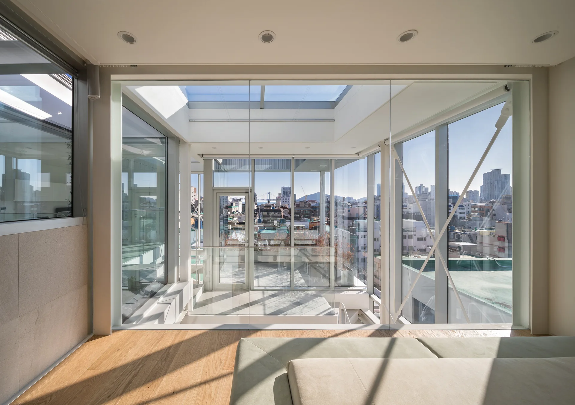A modern room with light wood floors features a green sofa looking through floor-to-ceiling glass walls and a skylight toward an urban cityscape.