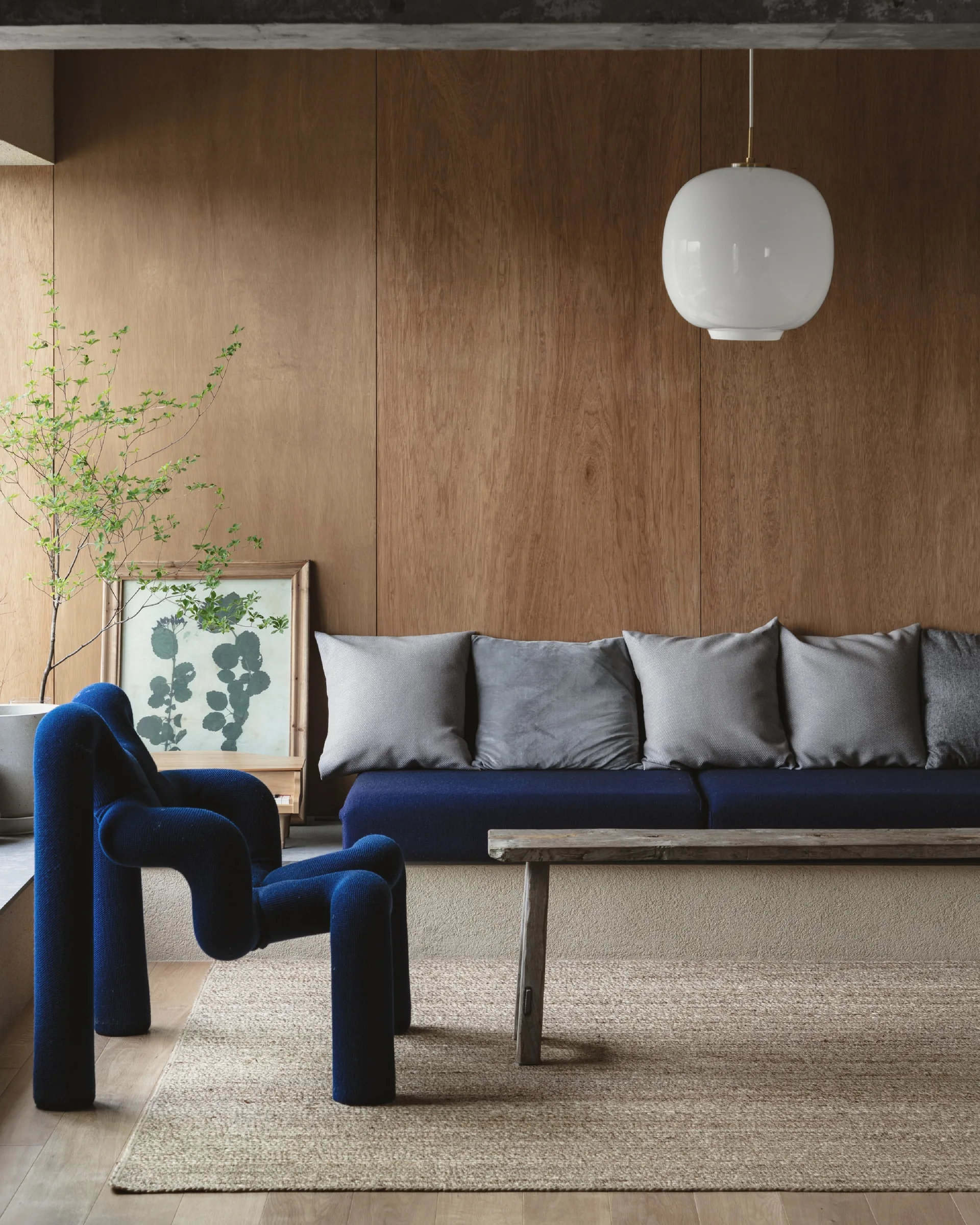 A modern living room features a built-in navy blue sofa and a sculptural blue armchair against warm wood-paneled walls, with a rustic wood bench.