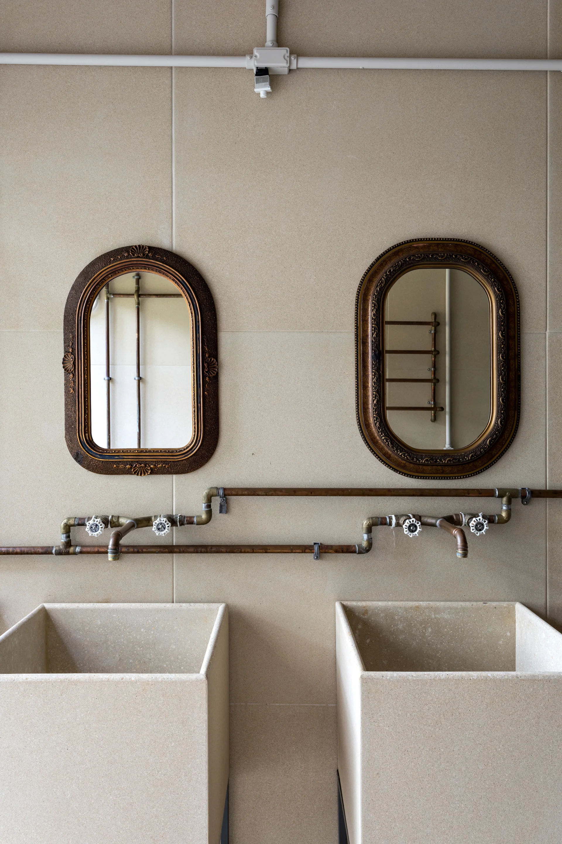 Two square stone basin sinks sit beneath ornate vintage mirrors on a beige tile wall with exposed copper plumbing.