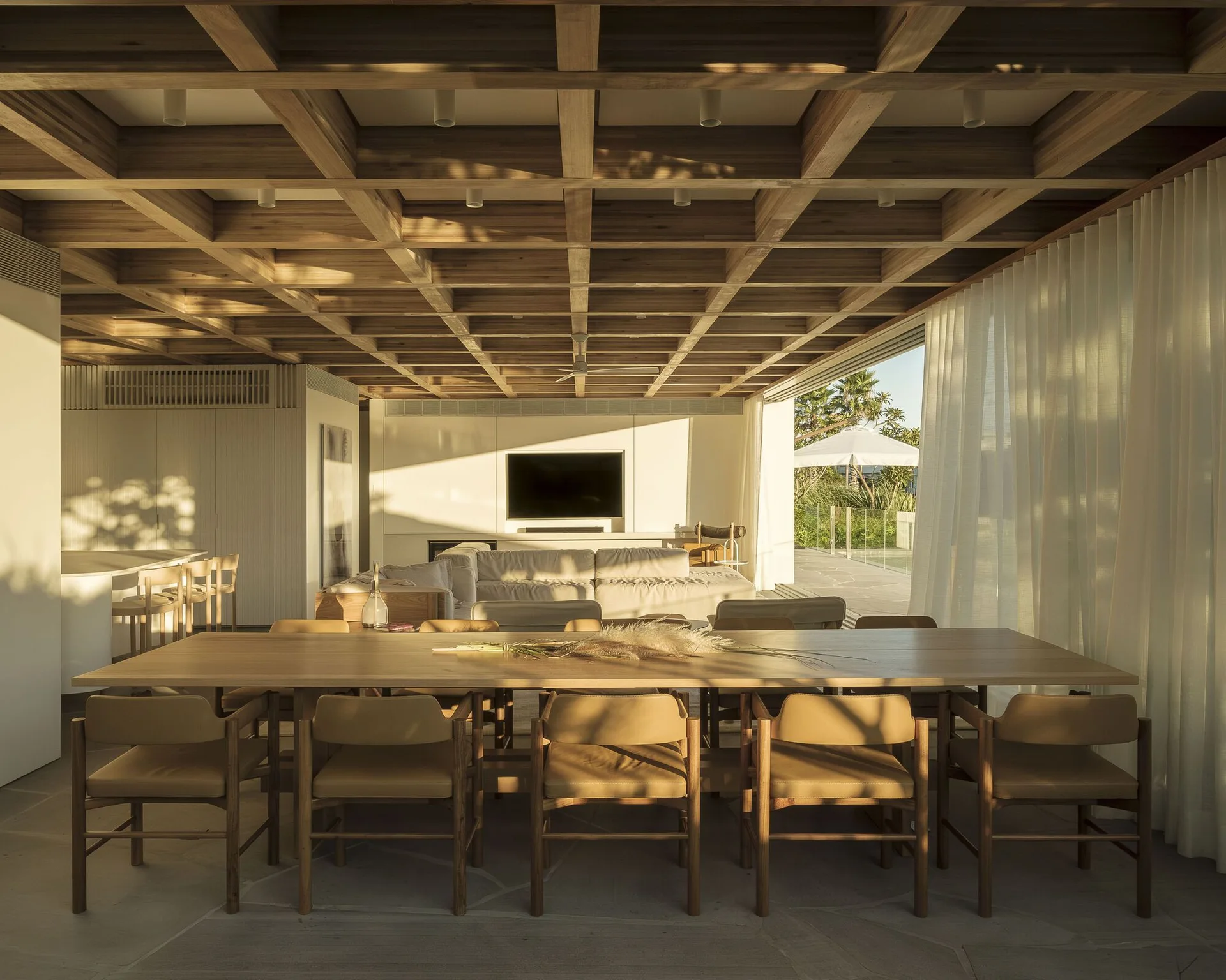 An open-plan dining room features a large wood table with tan chairs under a coffered wood ceiling next to a sunlit patio.