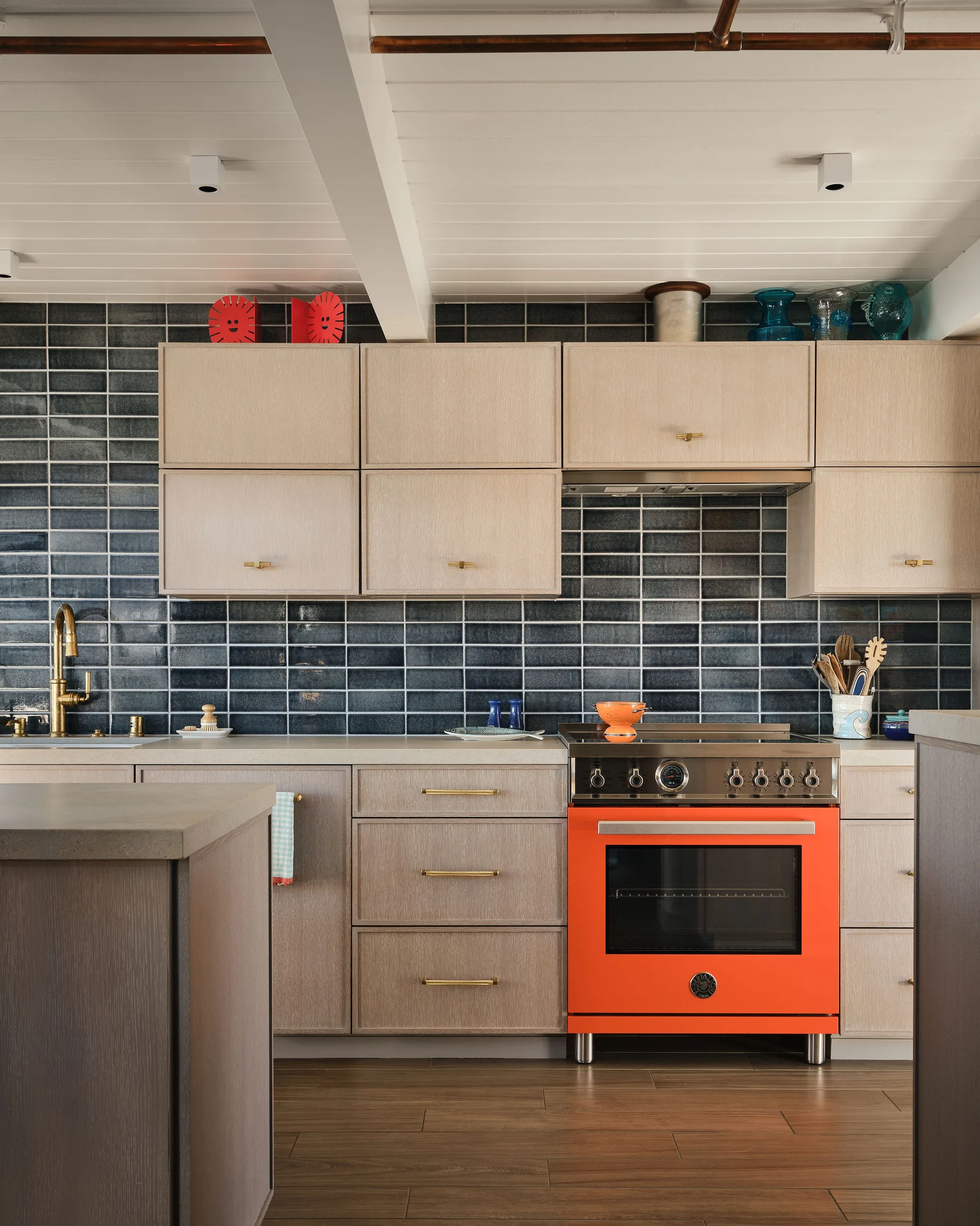 A modern kitchen features light wood cabinets, a glossy dark blue subway tile backsplash, and a bright orange range oven.