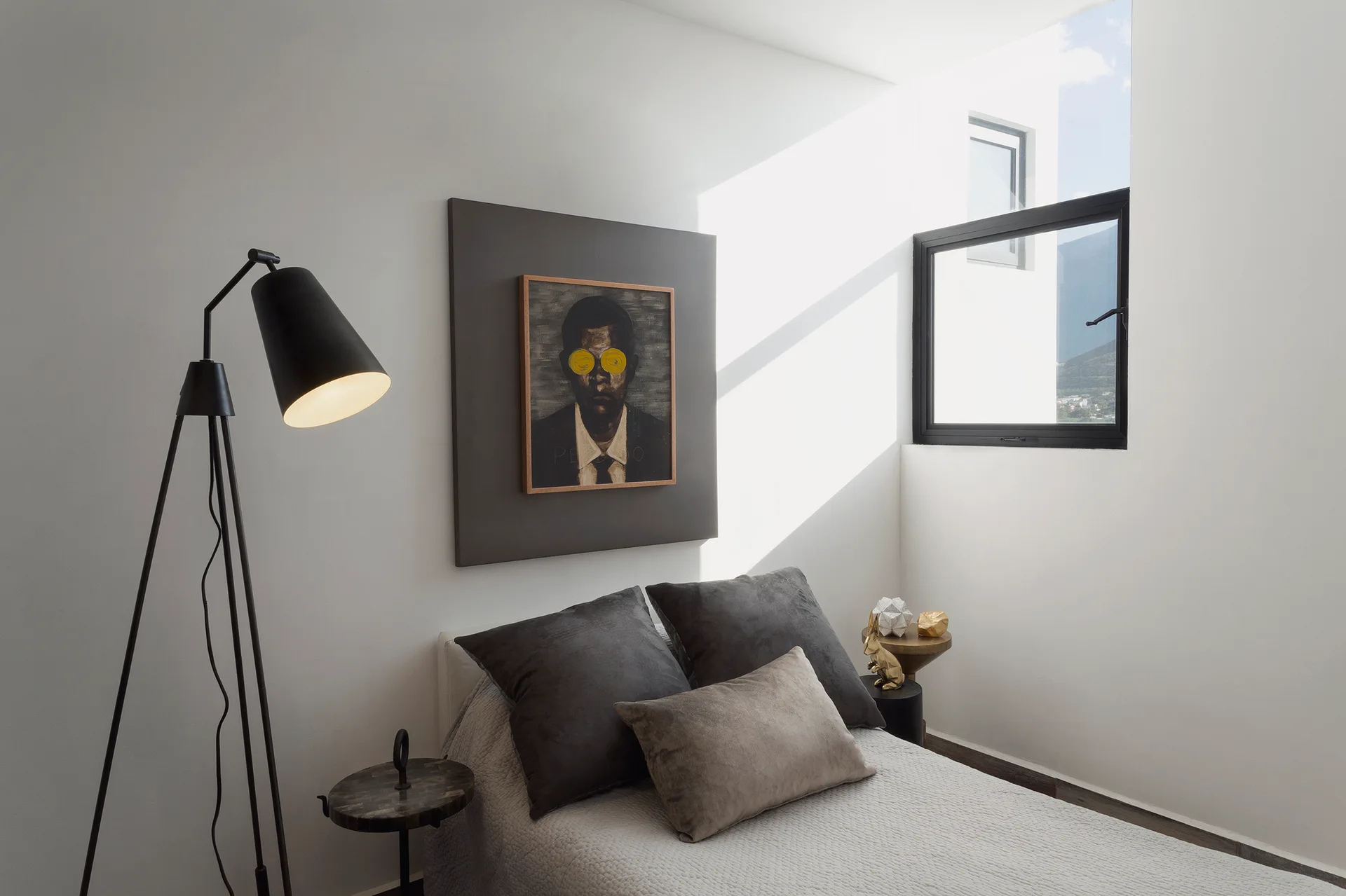 A minimalist bedroom features a bed with grey pillows, a black tripod floor lamp, and a portrait mounted on a large grey wall panel next to a window.