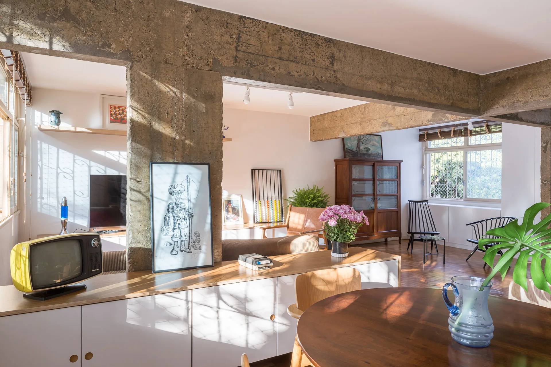An open-plan living and dining space featuring exposed concrete columns, vintage wood furniture, parquet flooring, and a low sideboard.