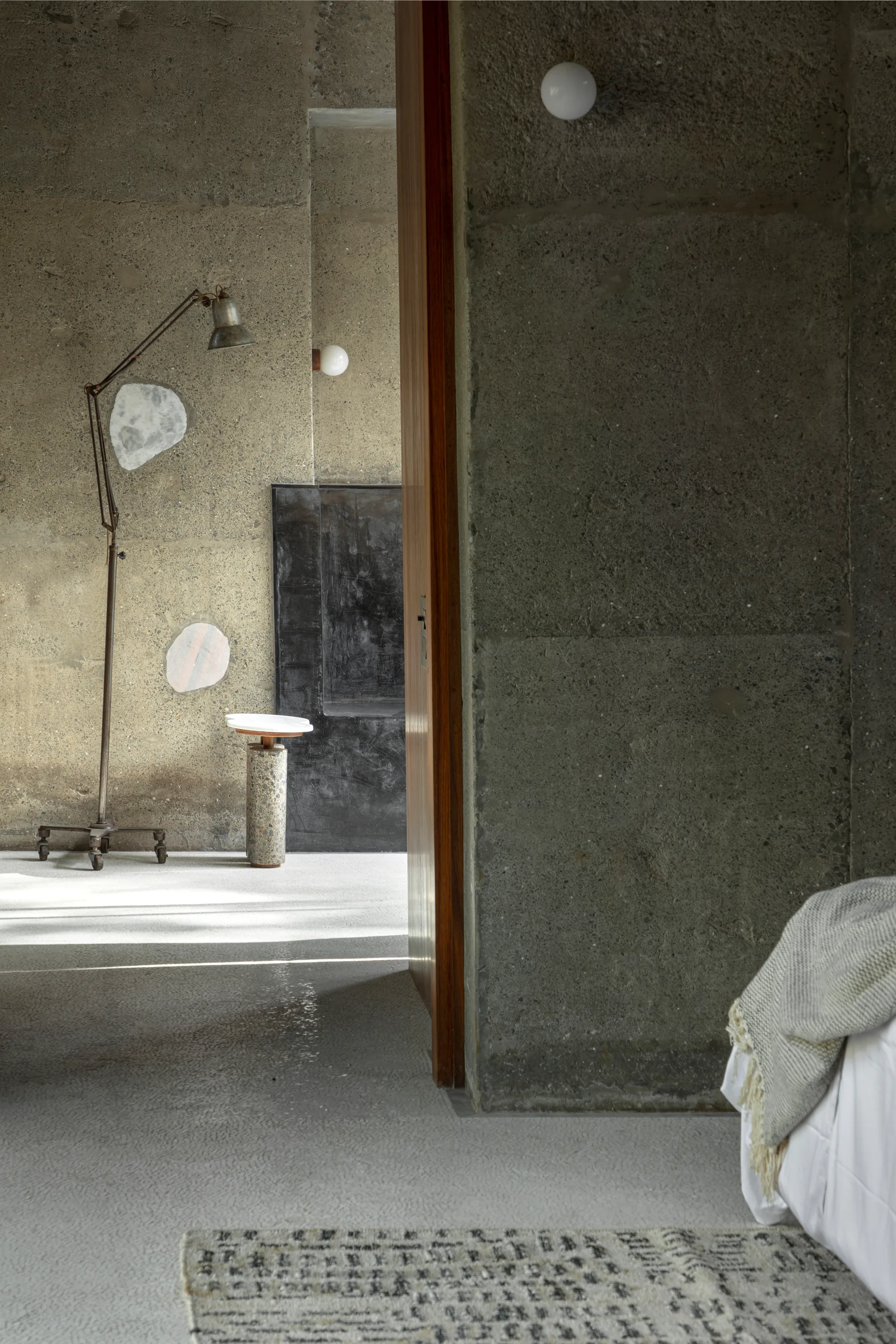 View through an open wooden door of a minimalist room with raw concrete walls, a vintage floor lamp, and a small stone pedestal table.