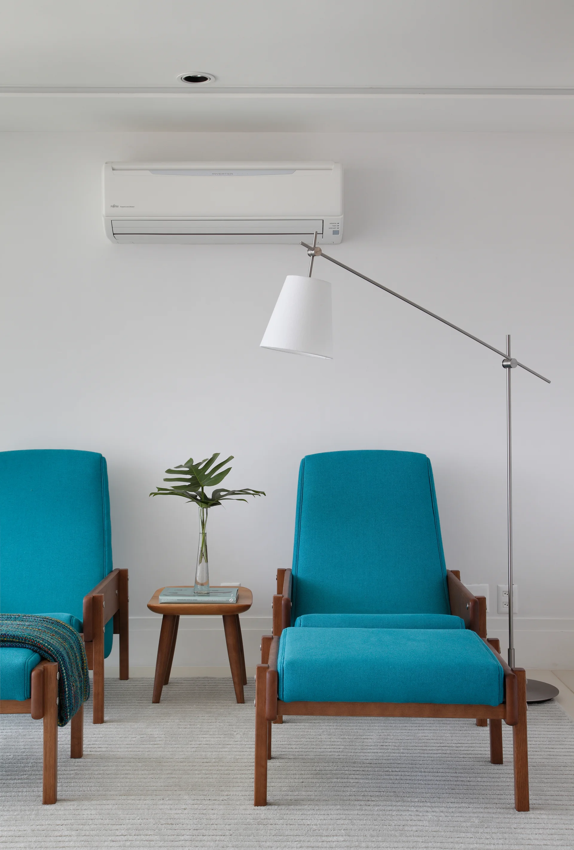 Two teal mid-century armchairs stand on a textured grey rug next to a small wooden side table and a silver floor lamp against a plain white wall.