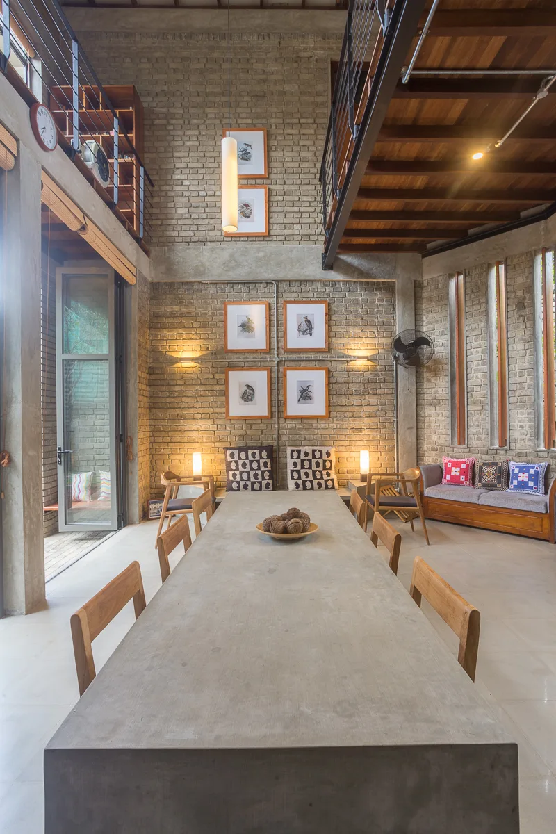 A dining room features a long concrete table, wooden chairs, and exposed brick walls beneath a wooden mezzanine.