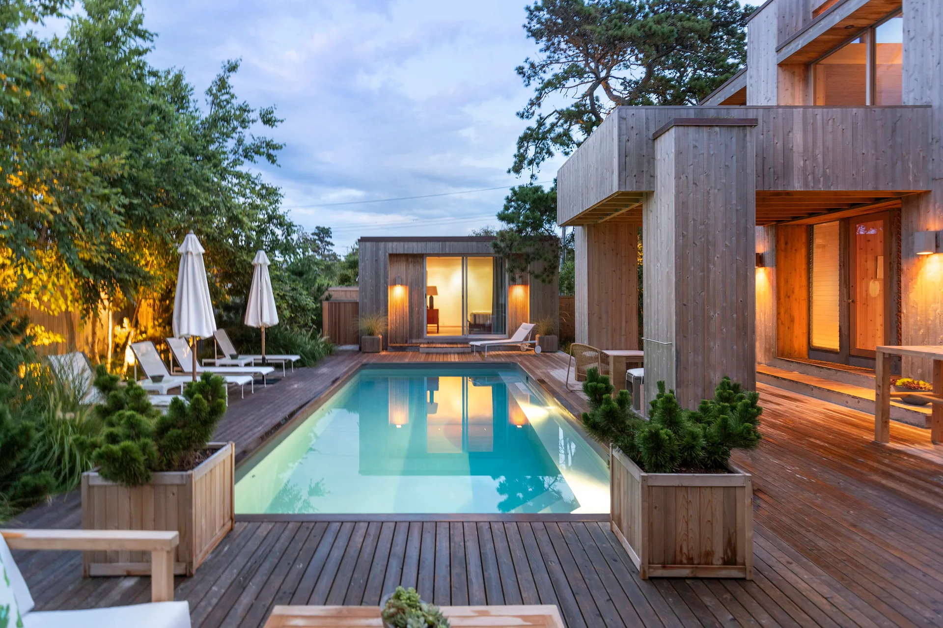 A rectangular swimming pool lit from within sits in a large wooden deck next to a modern wood-clad house at dusk.