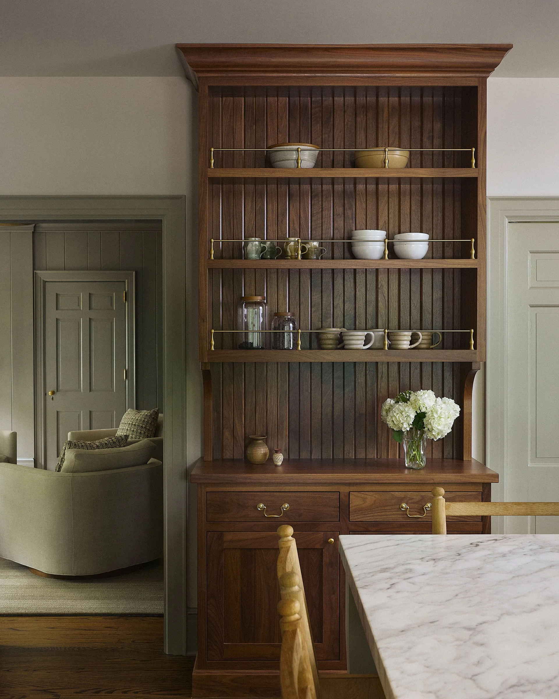 A dark wood built-in dining hutch with brass shelf rails holds ceramic bowls and glassware next to a marble table and an open doorway.