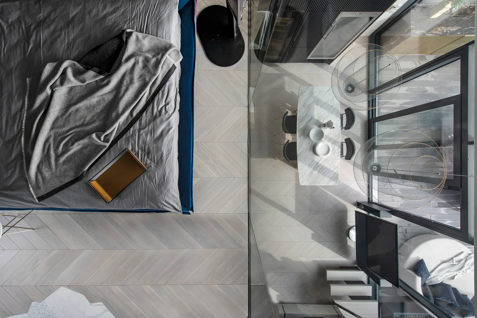 A top-down view of a modern loft bedroom with grey bedding overlooking a dining area with a marble table and herringbone wood floors.