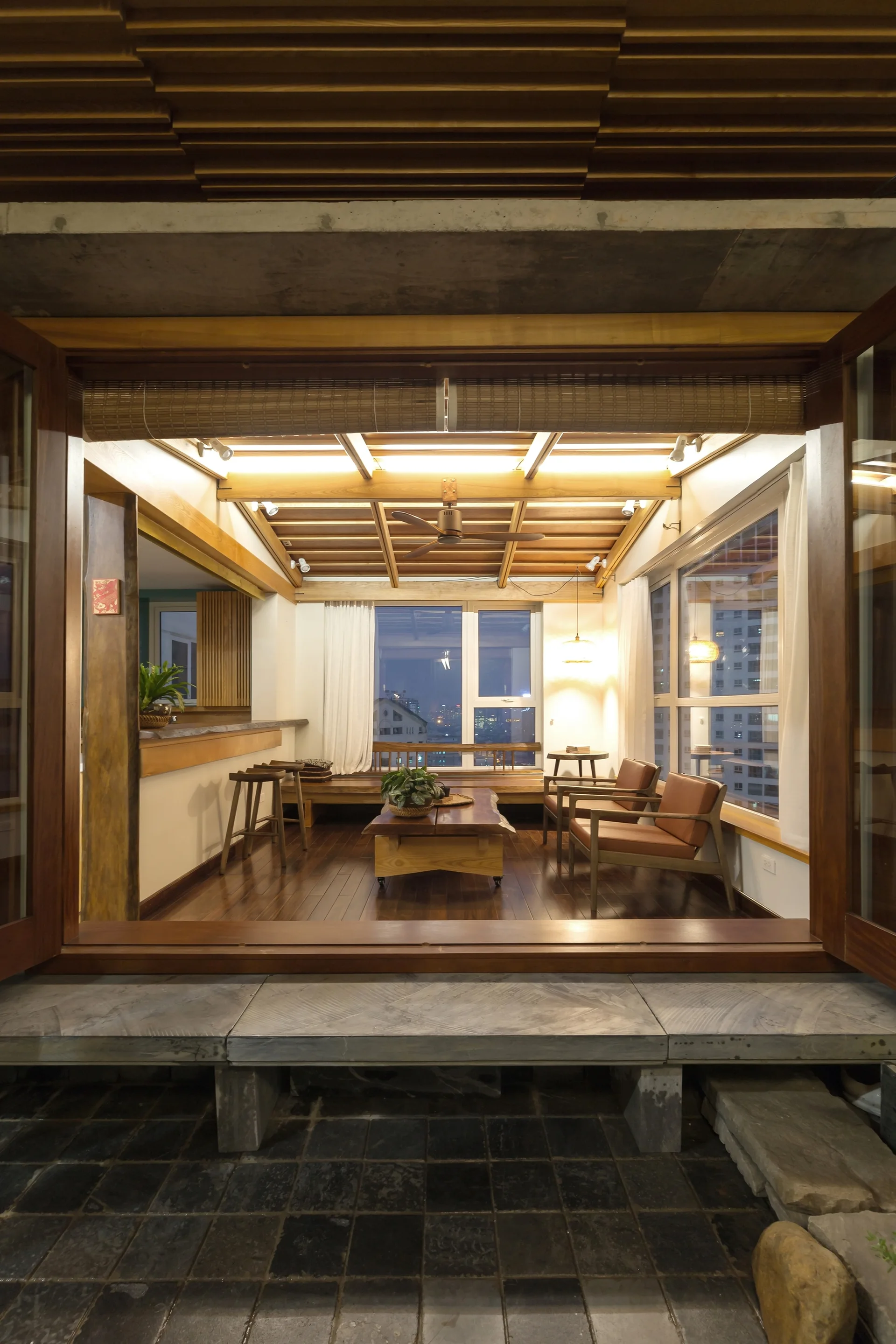 A cozy lounge area seen through wide doors features warm wooden ceilings, dark wood floors, a low live-edge table, and two leather armchairs facing windows.