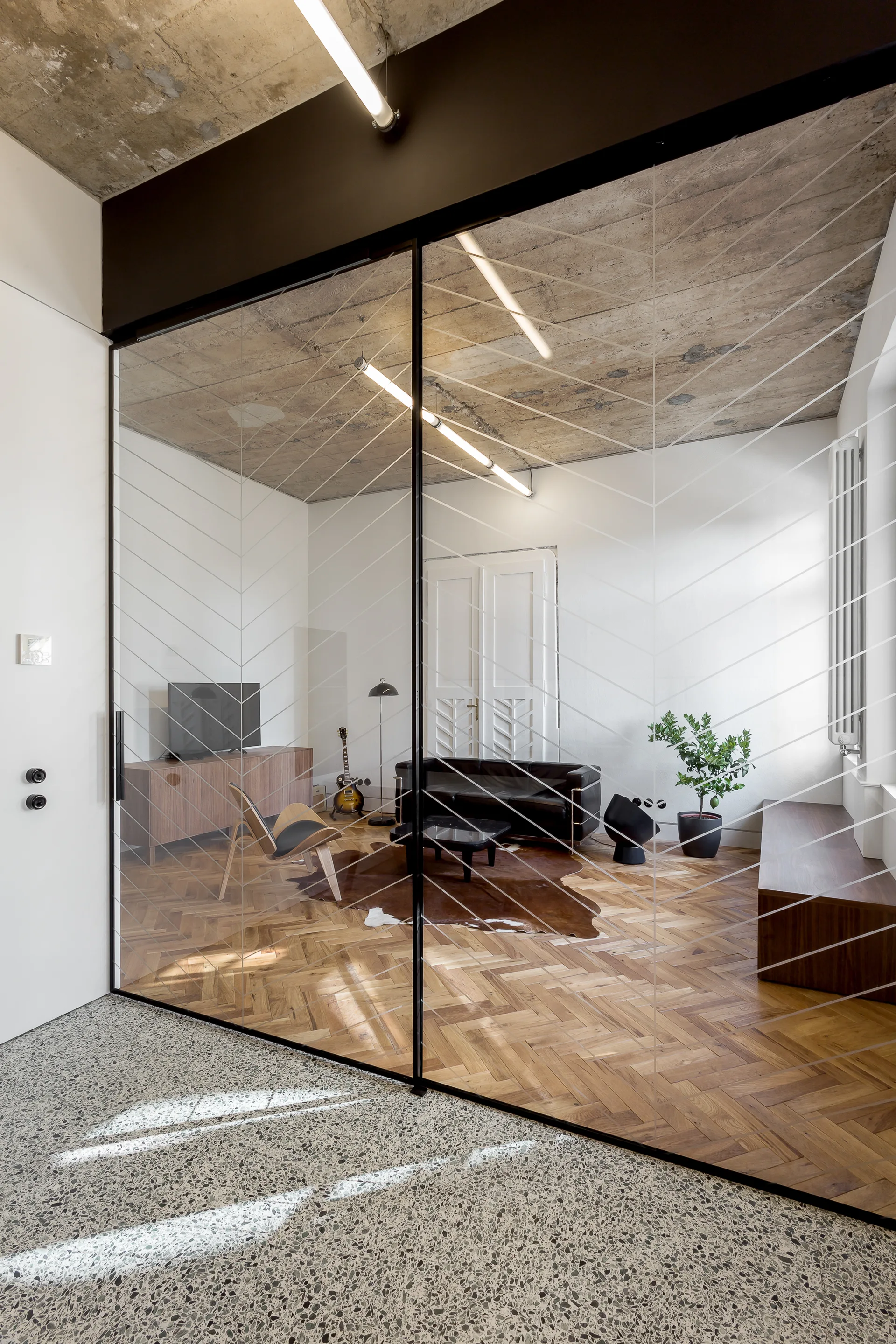 A living room with herringbone wood floors and a black leather sofa is viewed through a glass partition wall etched with chevron lines.