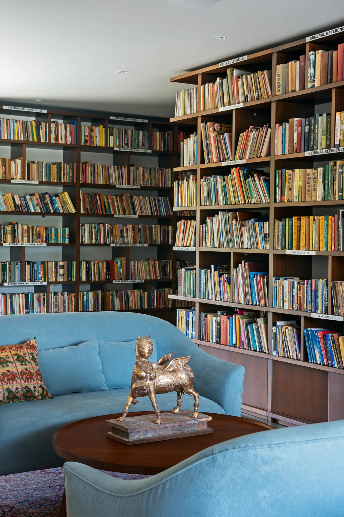 A home library featuring dark wood floor-to-ceiling bookshelves, light blue fabric sofas, and a round wooden coffee table with a gold sculpture.